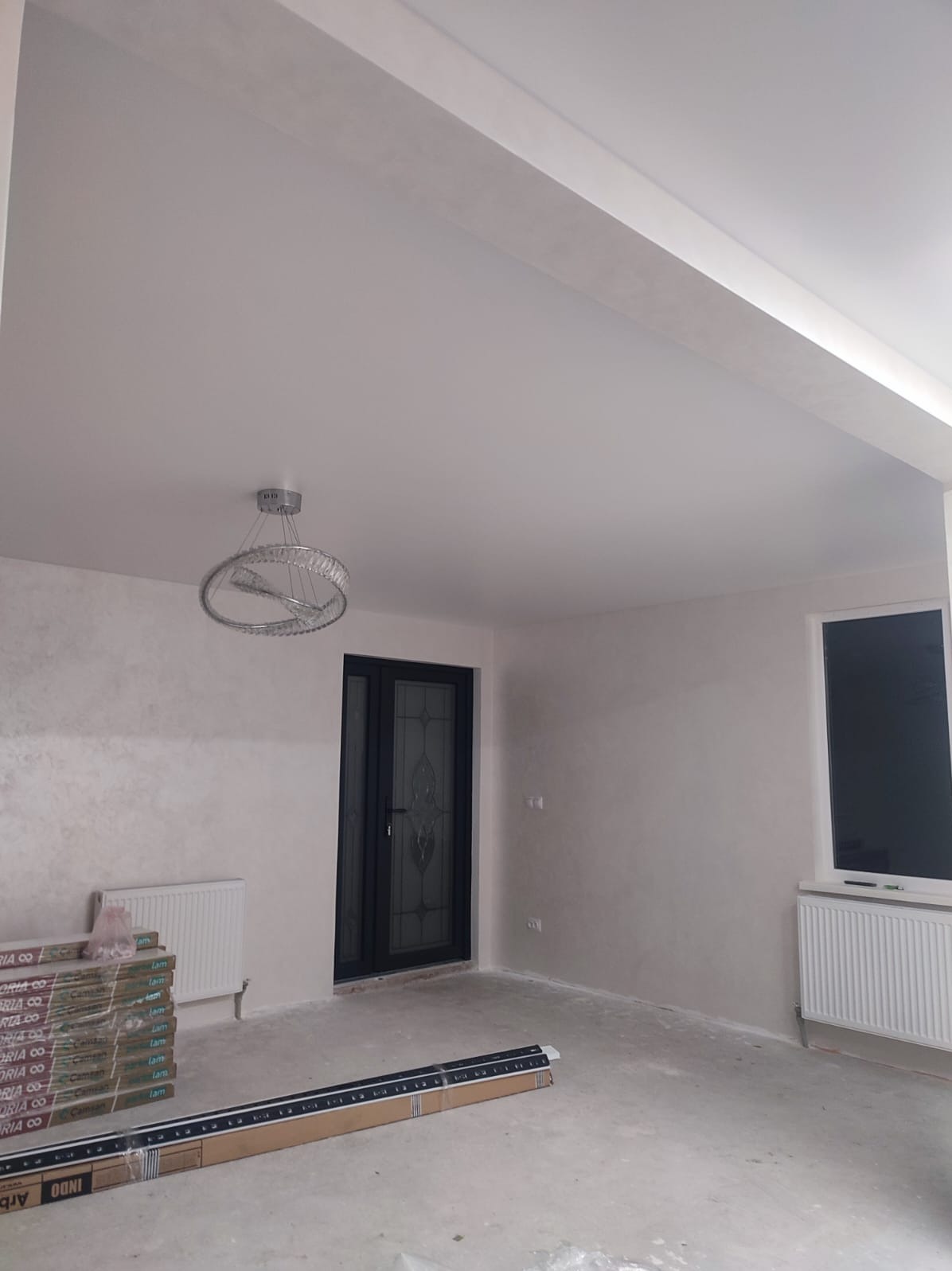 Wnętrze po gładziowaniu ścian i sufitu, z widoczną lampą, oknem, drzwiami i grzejnikiem. Na podłodze panele w opakowaniach. Ściany w jasnym odcieniu beżu.