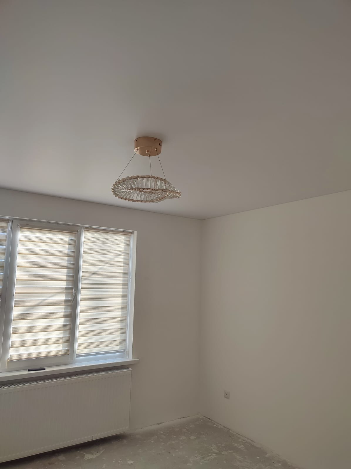 Jasne wnętrze po remoncie: świeżo pomalowane ściany w odcieniach bieli, okno z żaluzjami i ozdobny żyrandol. Podłoga przygotowana do dalszych prac wykończeniowych.