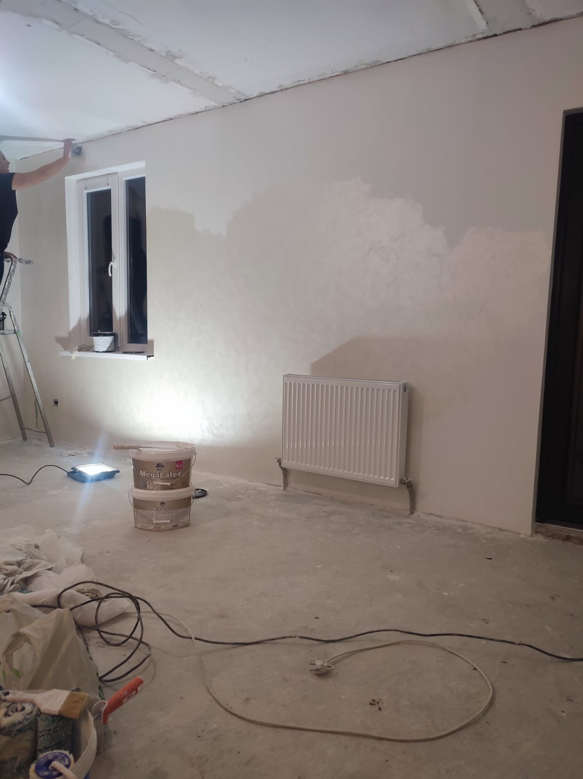 Wnętrze w trakcie malowania: ściana z grzejnikiem, okno, drabina i wiaderka z farbą Mega Latex. Widoczne niedomalowane fragmenty ściany i narzędzia malarskie.