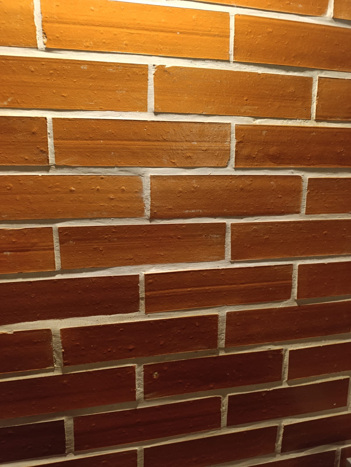 Ceglana ściana w odcieniach brązu z jasną fugą, ułożona w regularny wzór. Widoczne nierówności i tekstura cegieł, oświetlenie podkreśla detale powierzchni.
