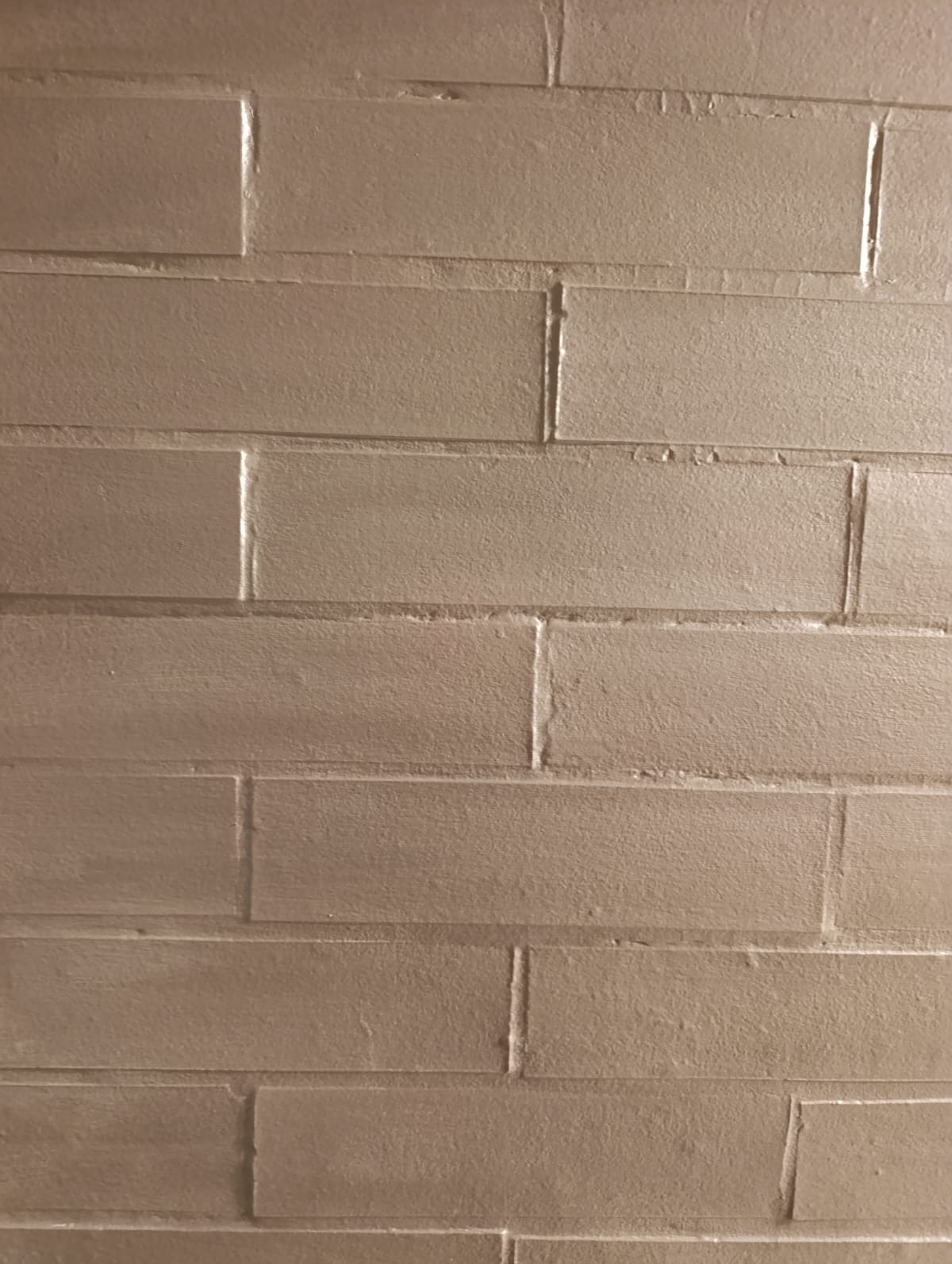 Teksturowana ściana pomalowana na jednolity, beżowy kolor, imitująca cegły. Widoczne nierówności i niedoskonałości w strukturze farby.