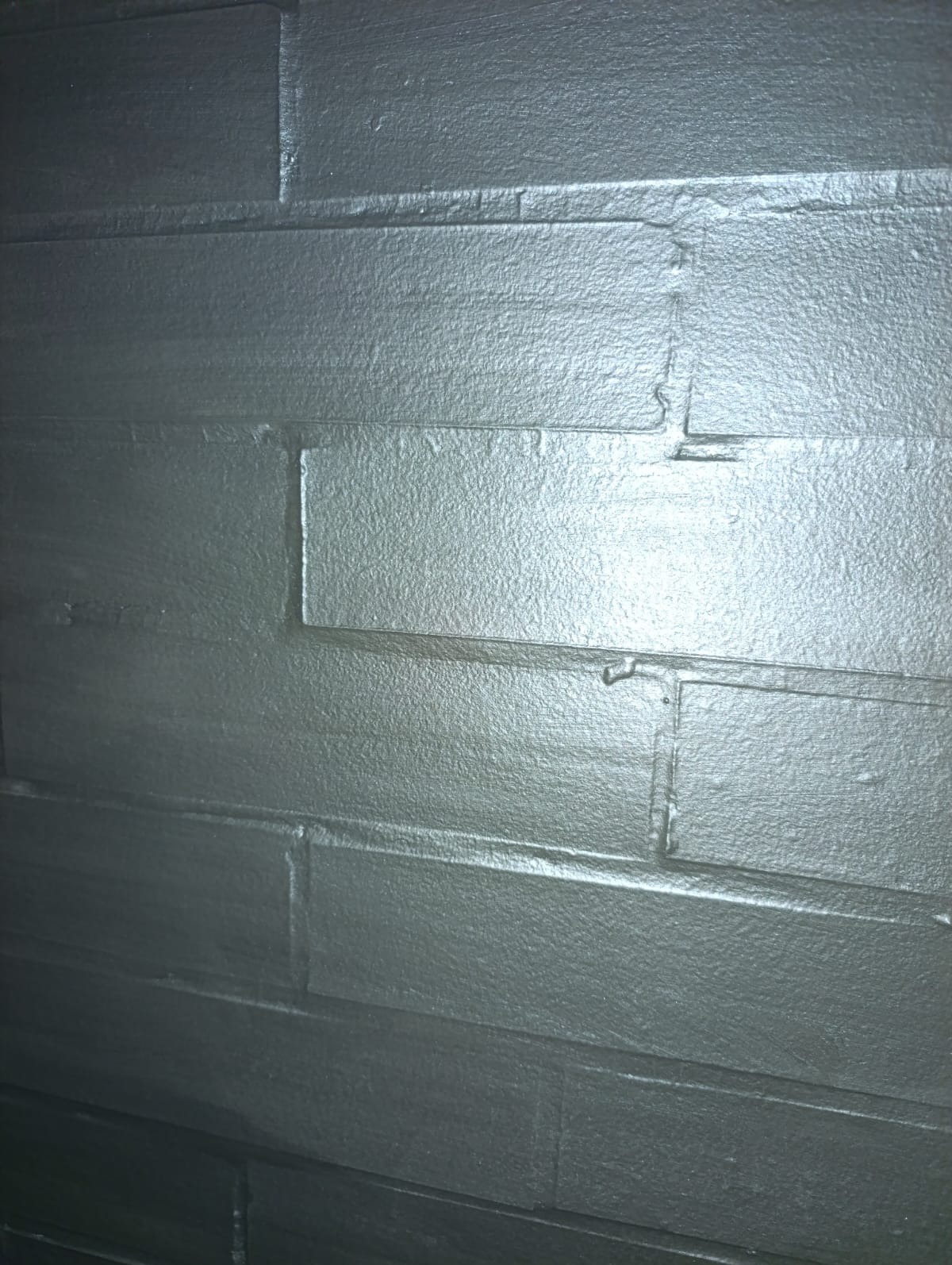 Ściana z cegieł pomalowana na srebrno, z widoczną fakturą i odbiciami światła, ukazująca detale wykończenia i nierówności powierzchni.