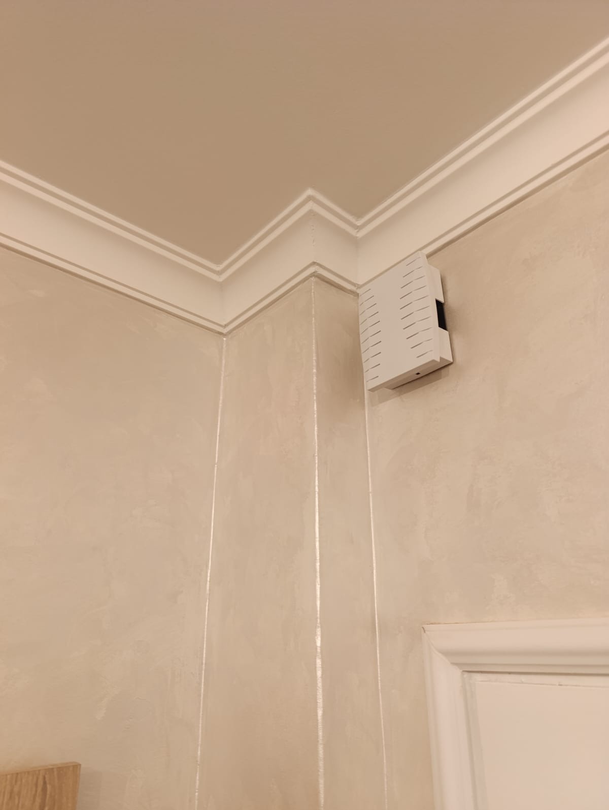 Beżowa ściana z dekoracyjną fakturą i białym gzymsem sufitowym. Widoczny fragment białej futryny drzwi i biała skrzynka elektryczna. Ujęcie z dołu, podkreślające detale wykończenia.