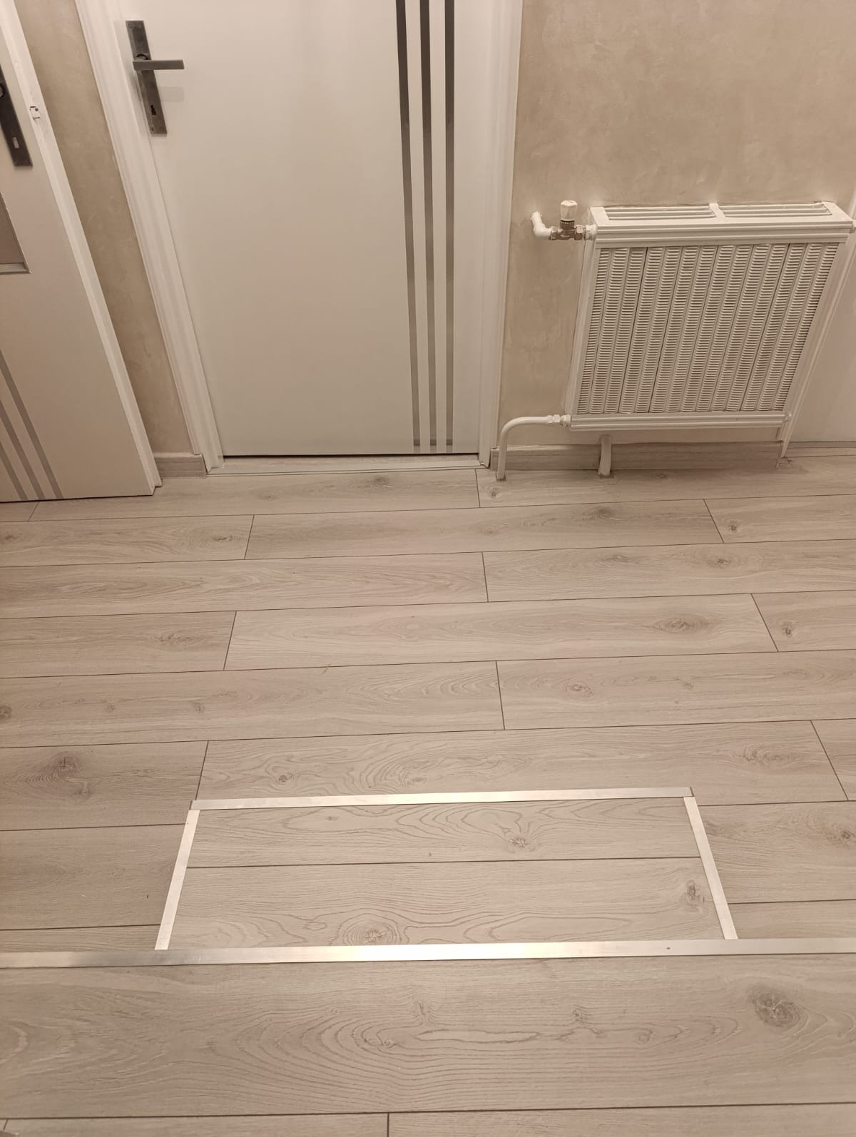 Jasne panele podłogowe z widocznym fragmentem maskownicy, biały grzejnik i drzwi w tle. Ujęcie z niskiej perspektywy.