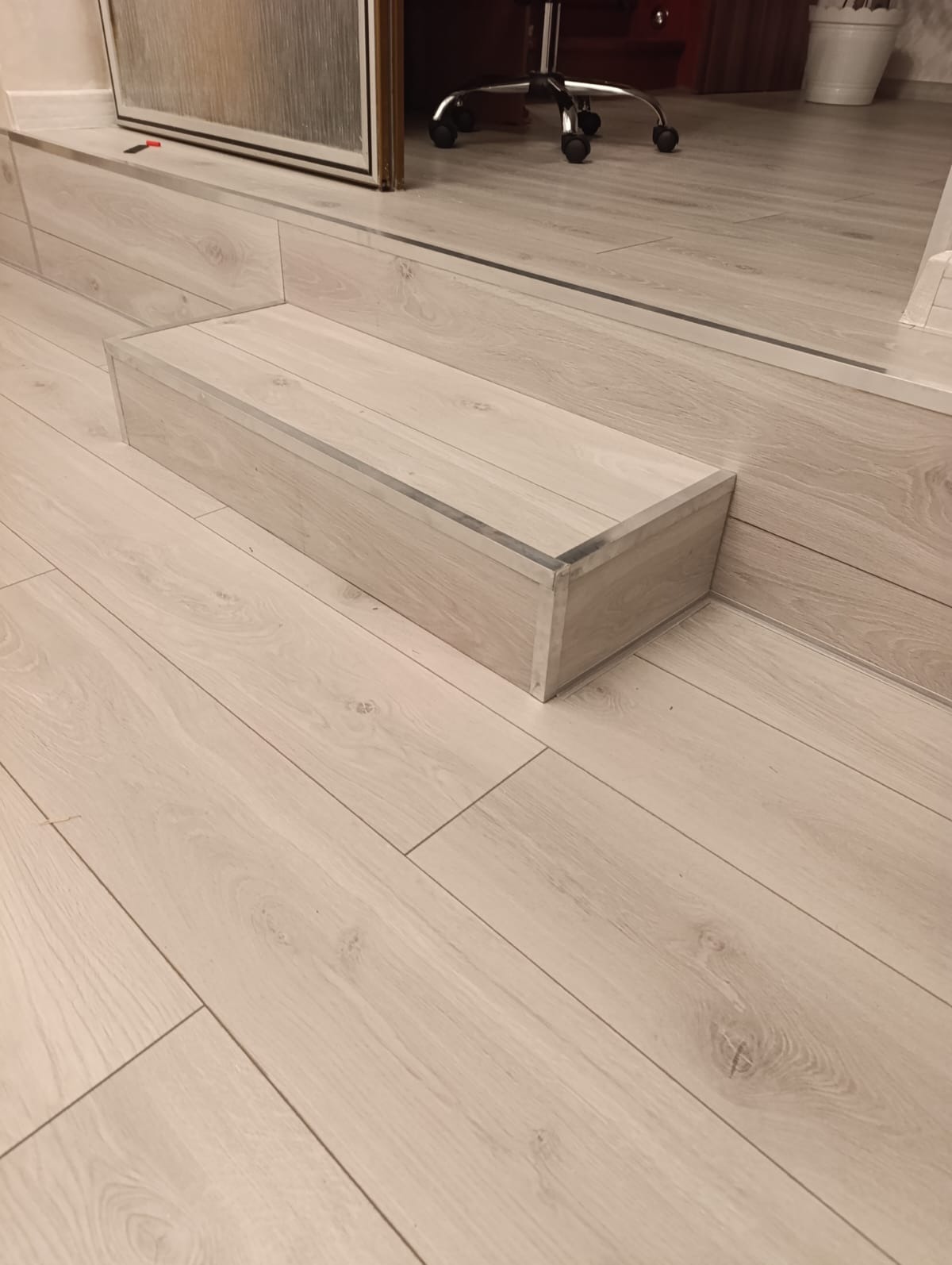 Jasne panele podłogowe z drewnopodobnym wzorem, widoczny stopień z listwą wykończeniową. W tle fragment biurowego krzesła i drzwi.