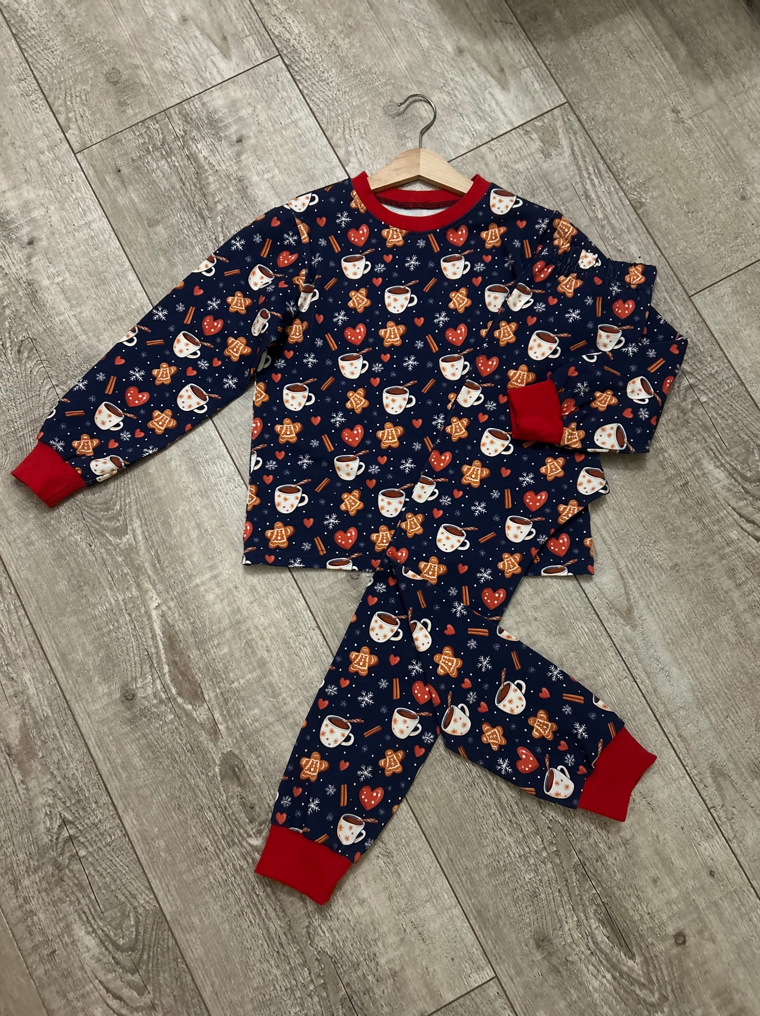 Świąteczna piżama dziecięca z motywem pierników i kubków z kakao, leżąca na drewnianej podłodze, zawieszona na drewnianym wieszaku. Czerwone wykończenia.