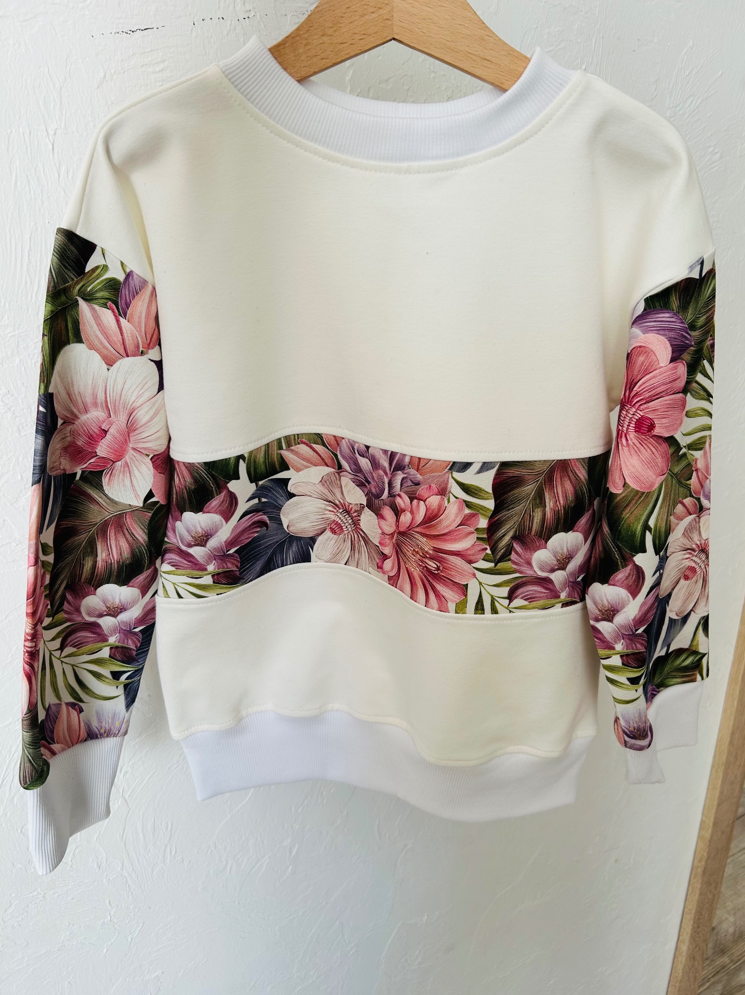 Kremowa bluza dziecięca z kwiatowym wzorem na rękawach i pasie, wisząca na drewnianym wieszaku na tle białej ściany. Widoczne detale wykończenia i jakość materiału.