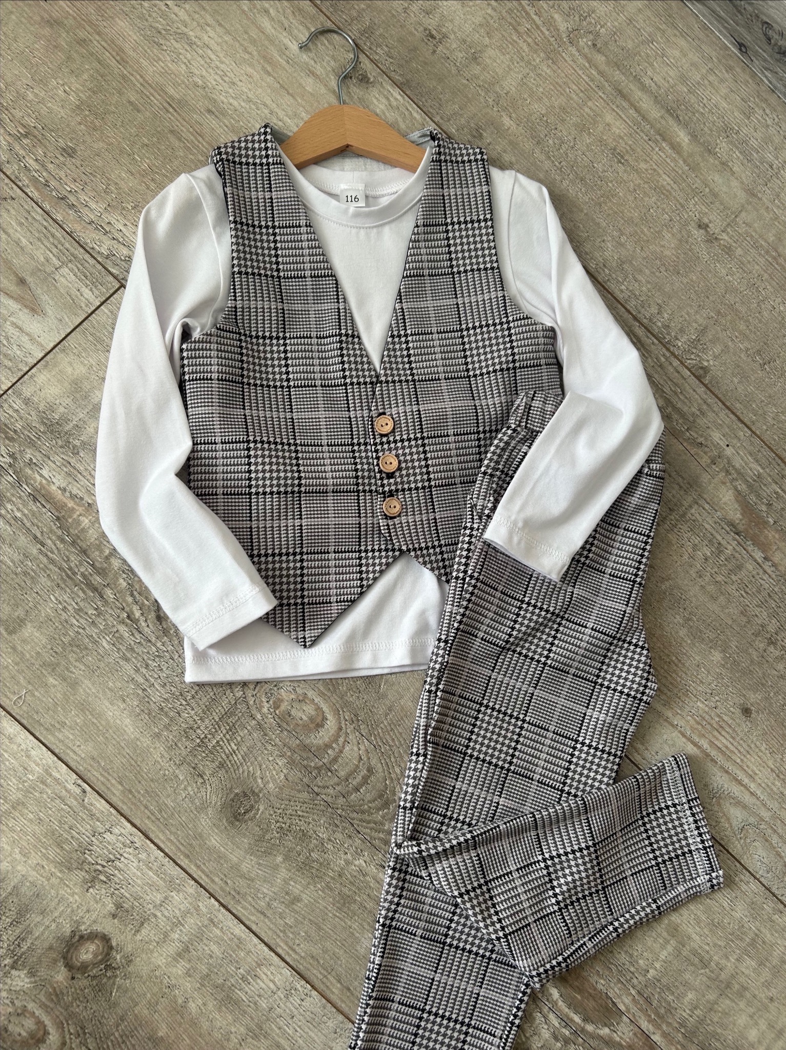 Elegancki komplet dziecięcy: biała bluzka z długim rękawem, kraciasta kamizelka z drewnianymi guzikami i spodnie w kratę, zawieszony na drewnianym wieszaku na tle drewnianej podłogi.