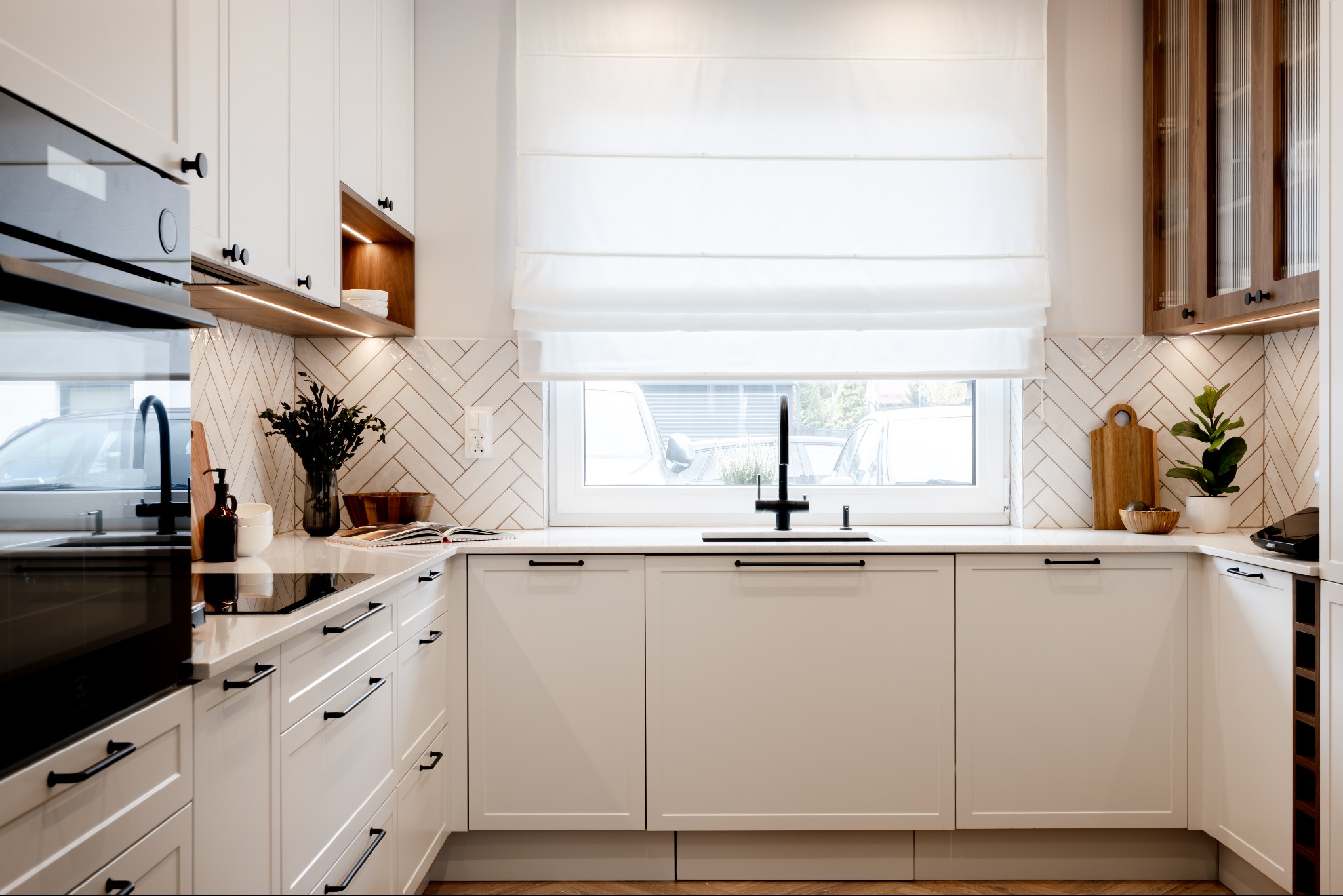 Jasna kuchnia z białymi szafkami, czarnymi uchwytami i armaturą. Na blacie dekoracje: deska do krojenia, roślina doniczkowa, wazon z kwiatami. Ściana wyłożona białą cegłą.