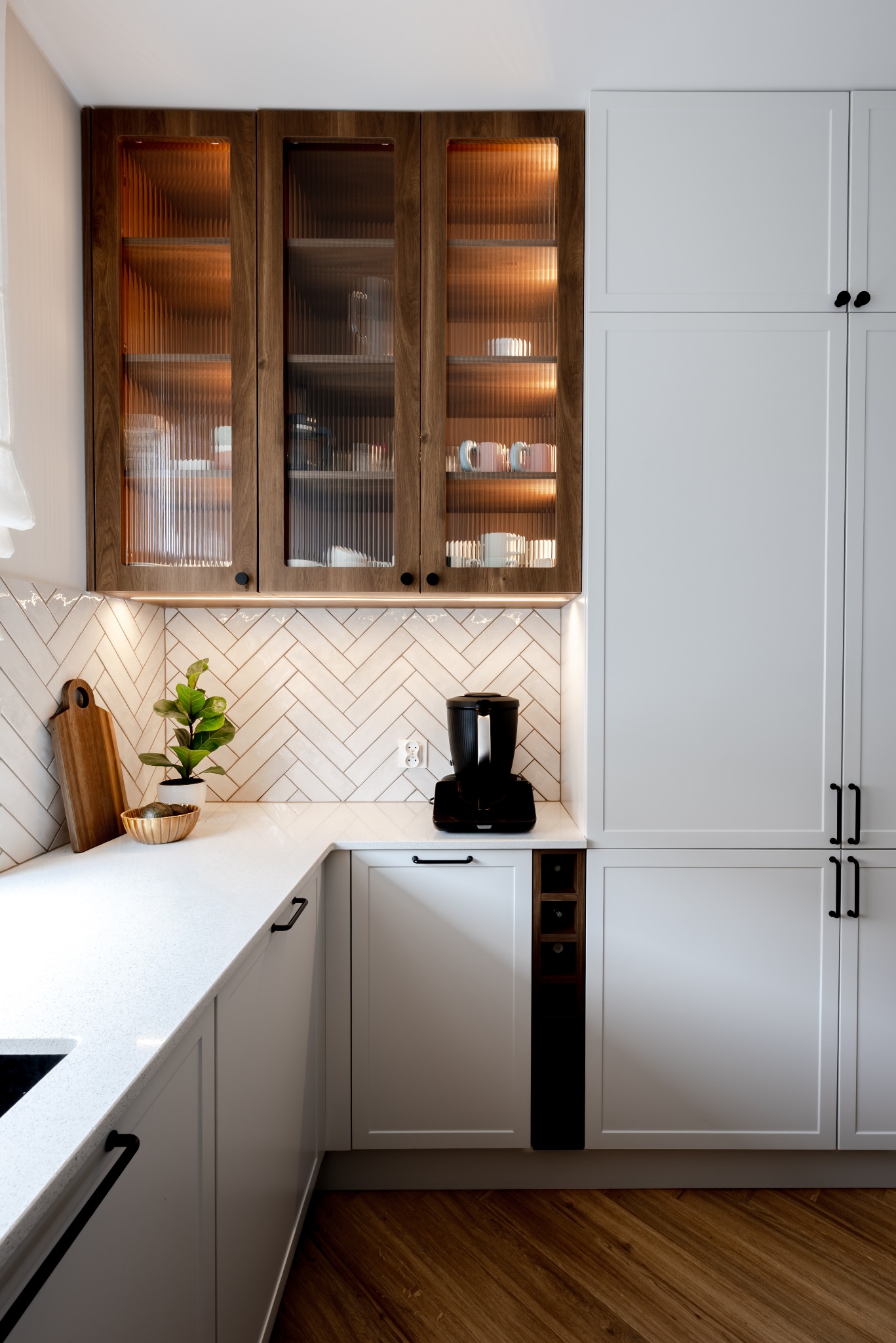 Nowoczesna kuchnia z białymi szafkami i drewnianymi akcentami. Na blacie deska do krojenia, roślina w złotej doniczce i czarny ekspres do kawy. Uwagę przykuwają ryflowane szklane fronty.