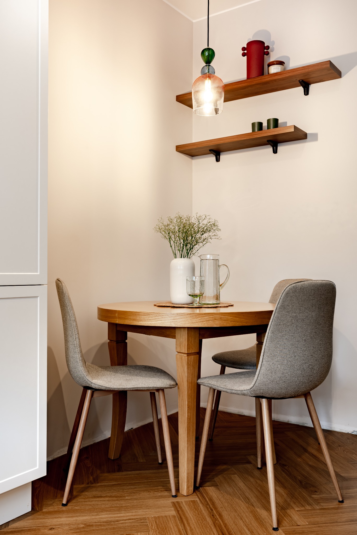 Jasne wnętrze: okrągły stół z drewna, szare krzesła, wazon z gipsówką, szklany dzbanek. Na ścianie półki z dekoracjami i lampa wisząca.