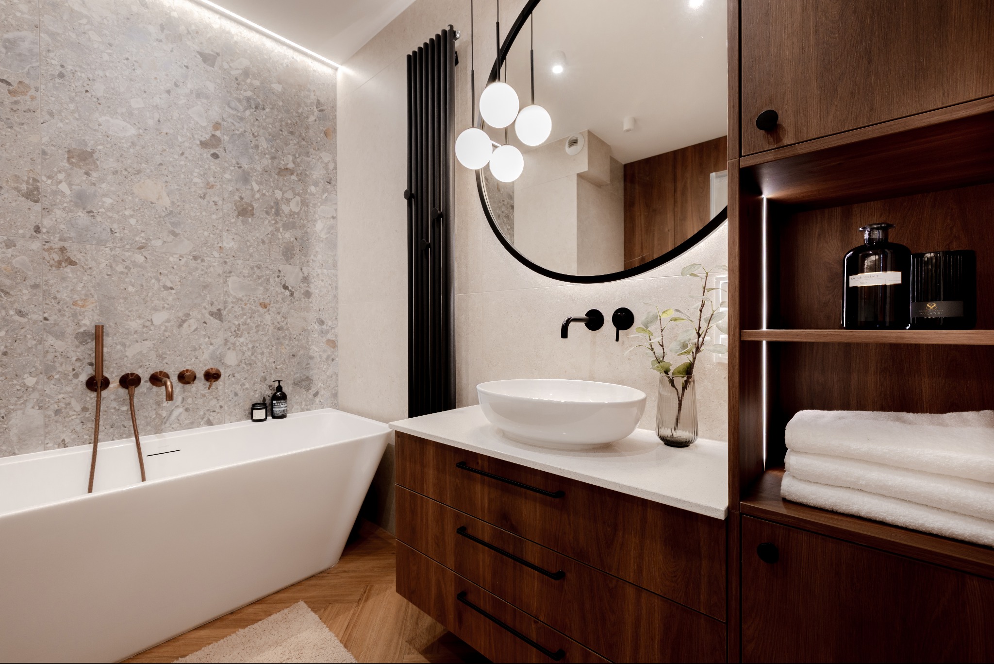 Nowoczesna łazienka z wolnostojącą wanną, umywalką nablatową i drewnianymi szafkami. Uwagę zwraca okrągłe lustro z oświetleniem i designerski grzejnik.