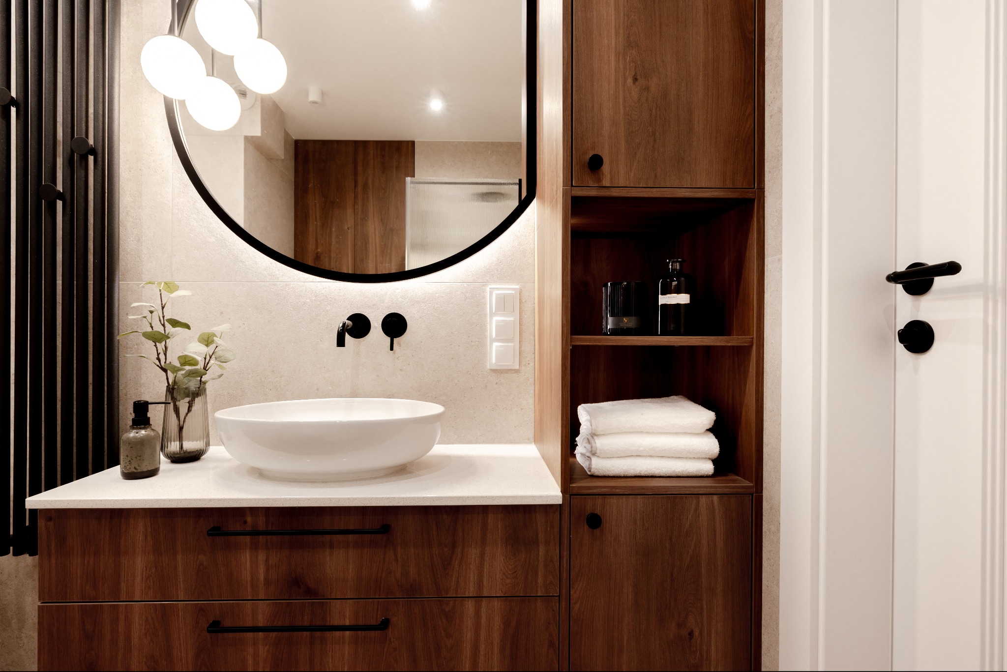 Nowoczesna łazienka z okrągłym lustrem podświetlanym LED, drewnianymi szafkami i białą umywalką nablatową. Minimalistyczny design z czarnymi akcentami.