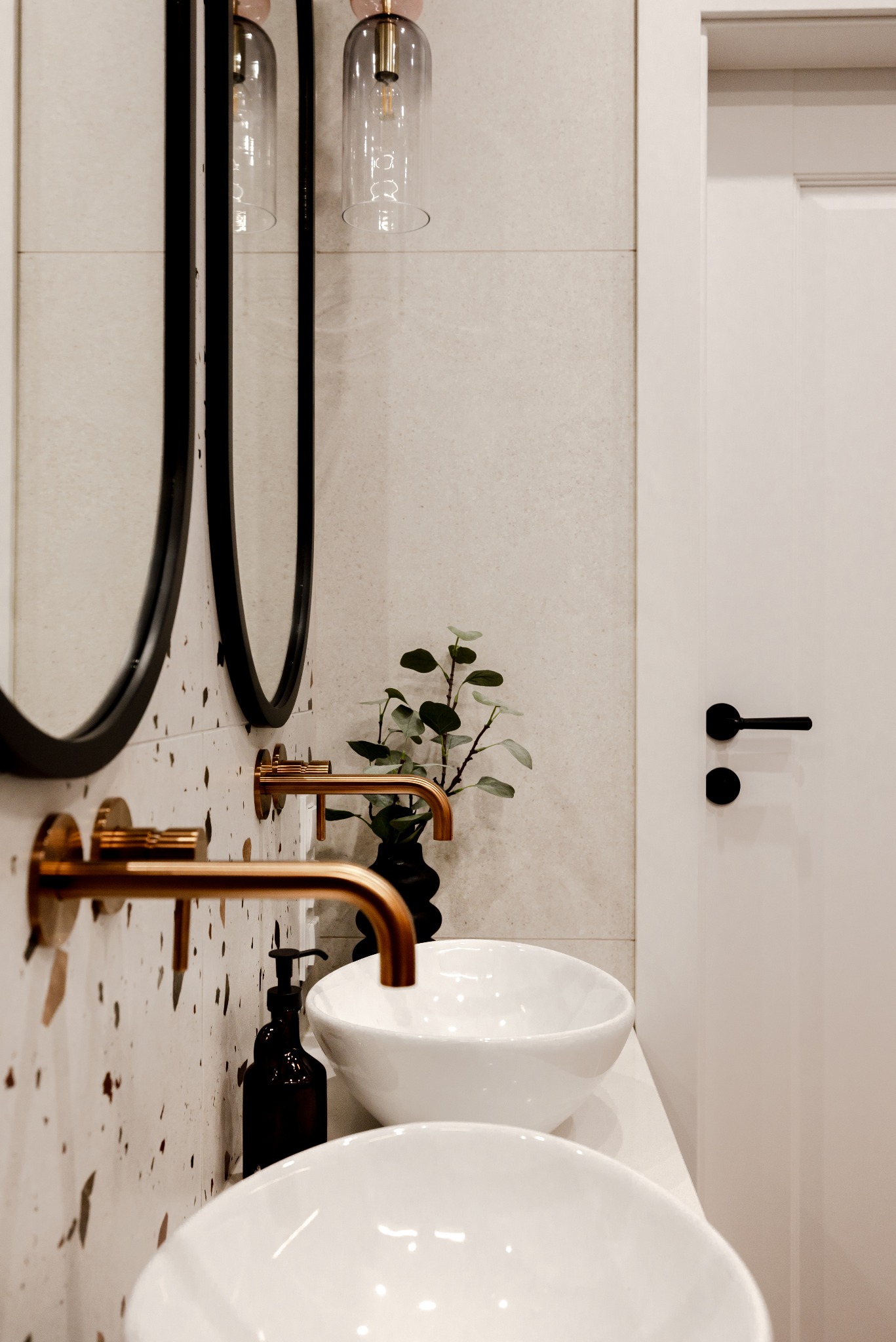Nowoczesna łazienka z dwoma białymi umywalkami, złotymi bateriami i okrągłymi lustrami na tle ściany w beżowe plamki. Minimalistyczny design z rośliną w czarnym wazonie.