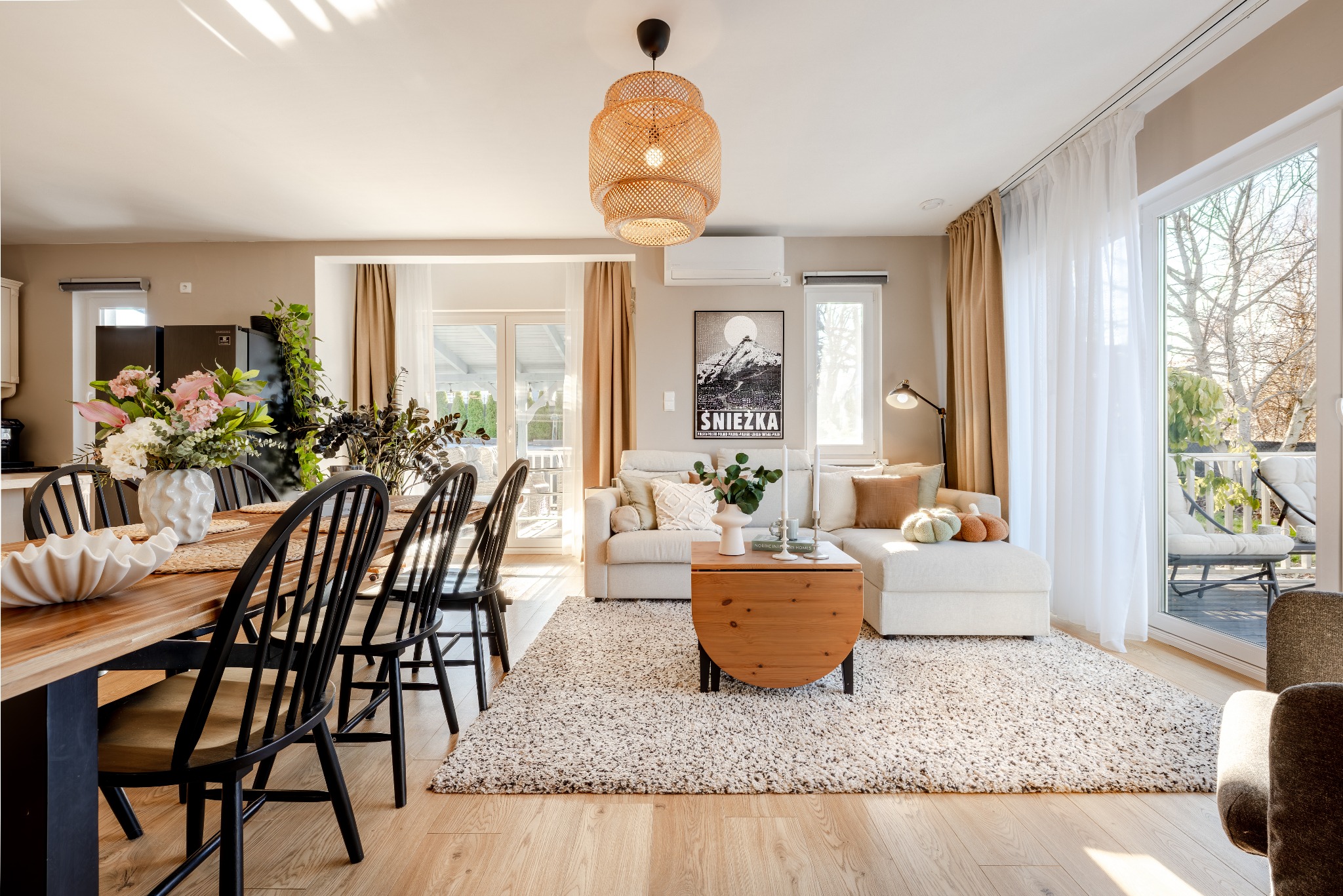 Jasne wnętrze z drewnianym stołem, czarnymi krzesłami i beżową sofą. W tle plakat z motywem górskim i balkon z widokiem na zieleń. Naturalne światło.