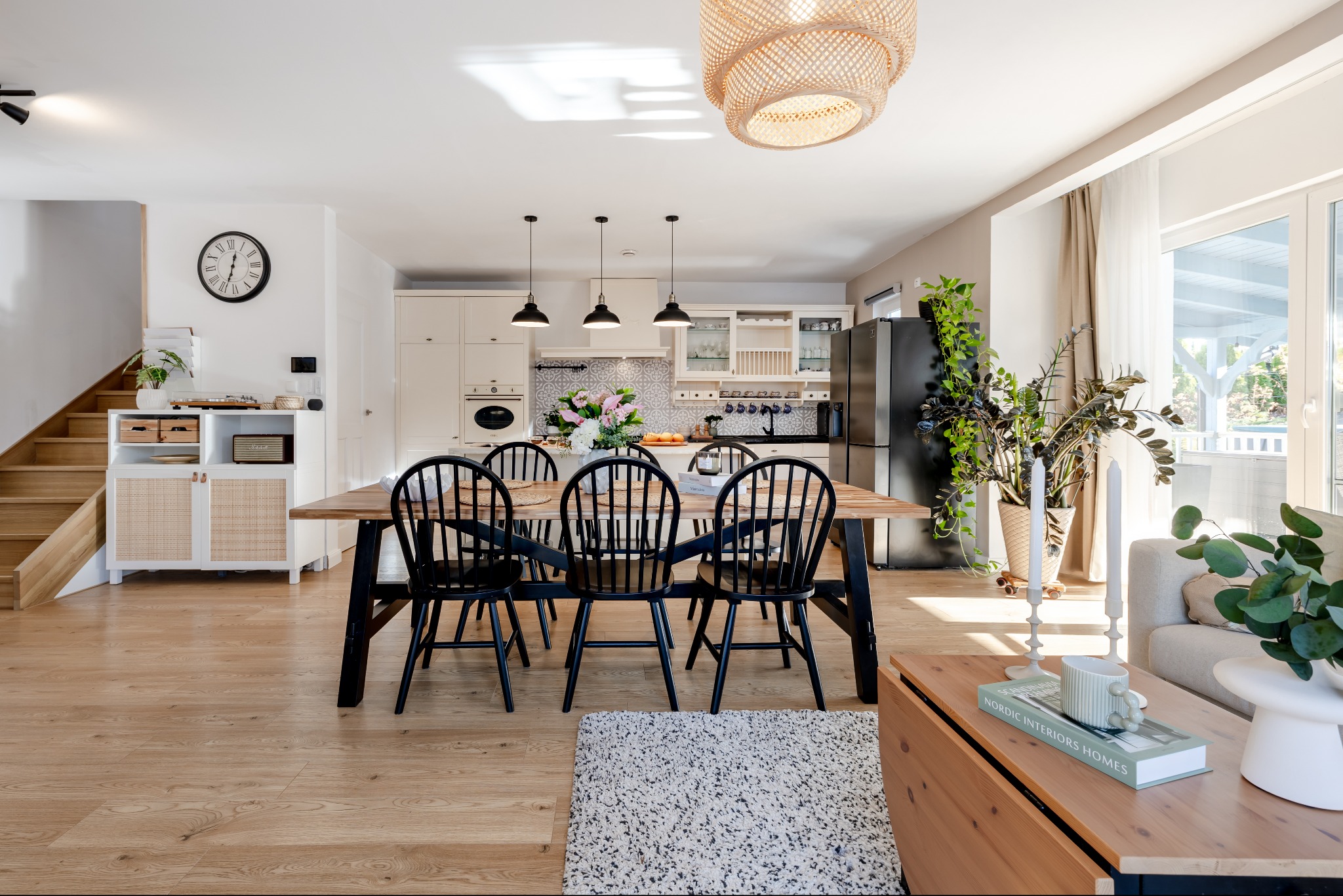 Jasne wnętrze z drewnianym stołem i czarnymi krzesłami. W tle widoczna kuchnia i schody. Całość dopełniają dekoracje roślinne i dywan.
