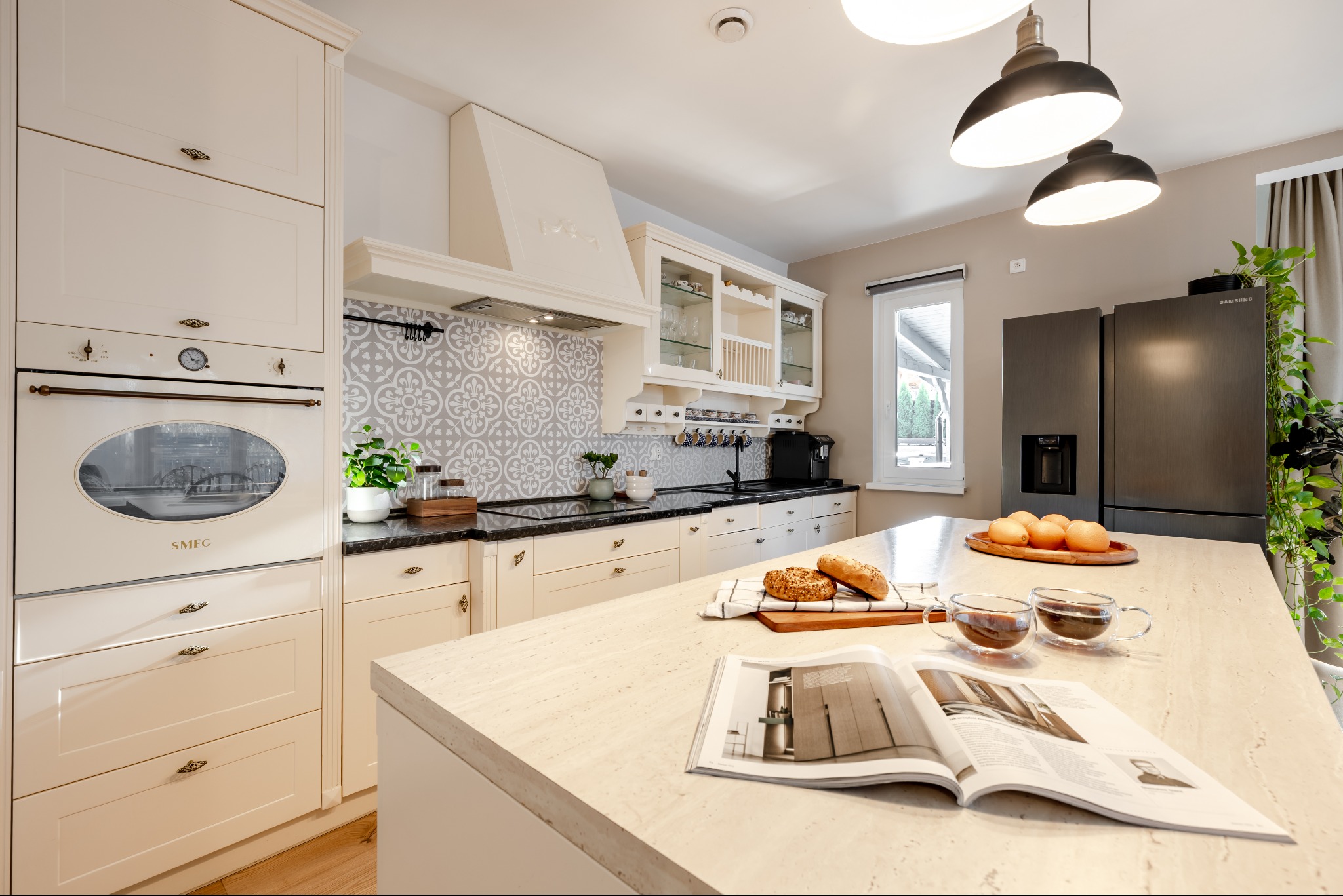 Jasna kuchnia w stylu retro z wyspą, na której leżą kawa, pieczywo i otwarta książka. Widoczny piekarnik SMEG i lodówka Samsung. Aranżacja wnętrz.