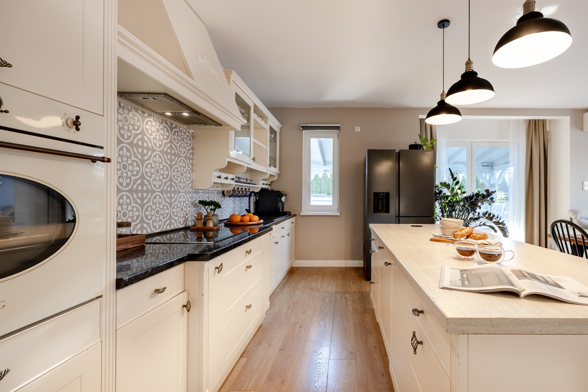 Jasna kuchnia w stylu retro z wyspą, czarnym blatem i wzorzystą mozaiką nad blatem. Na wyspie śniadanie i otwarta książka. Oświetlenie: trzy czarne lampy.