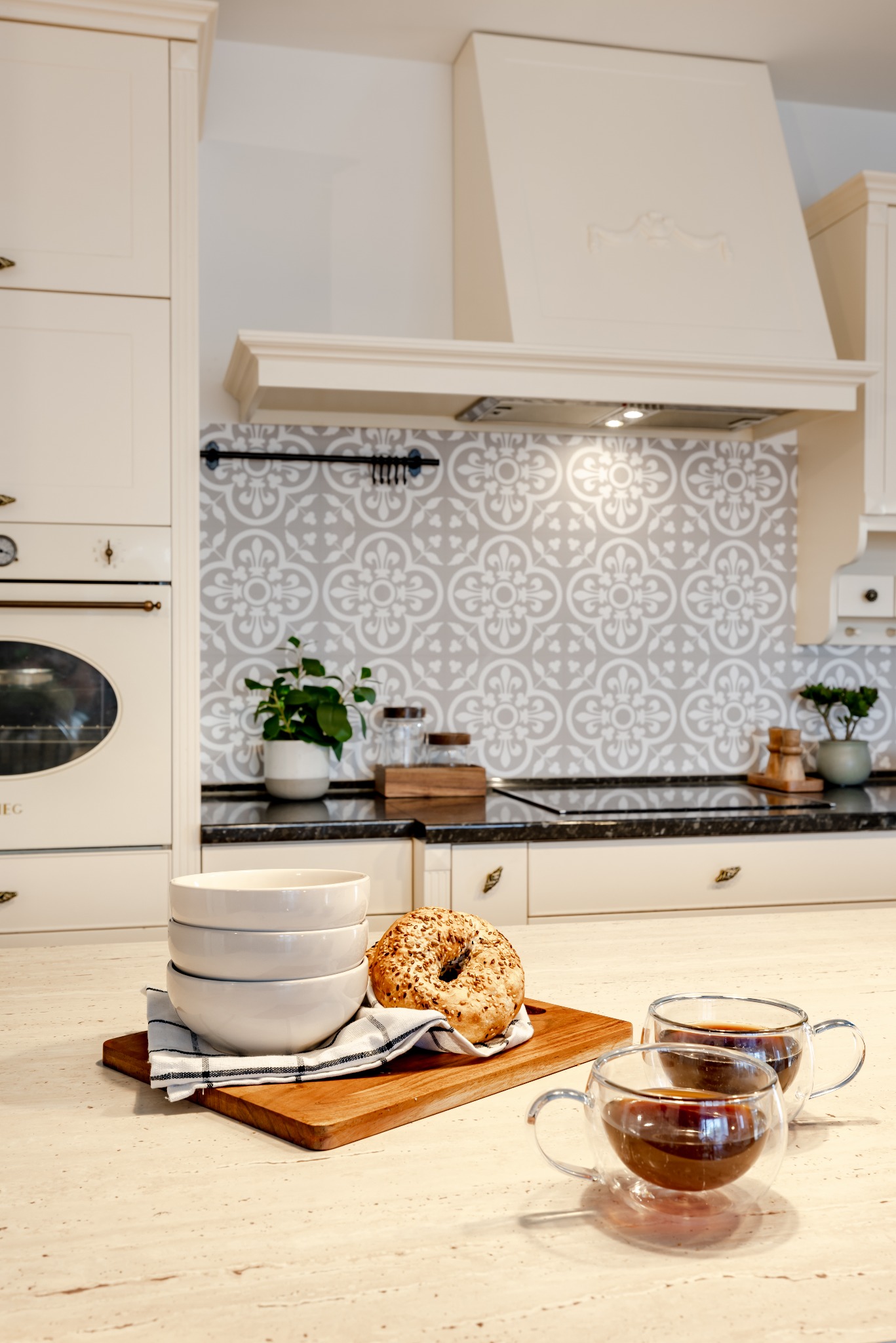 Jasna kuchnia w stylu retro z dekoracyjną płytką nad blatem. Na blacie drewniana deska z bułką, miskami i dwiema szklankami z kawą. Ciepłe oświetlenie.