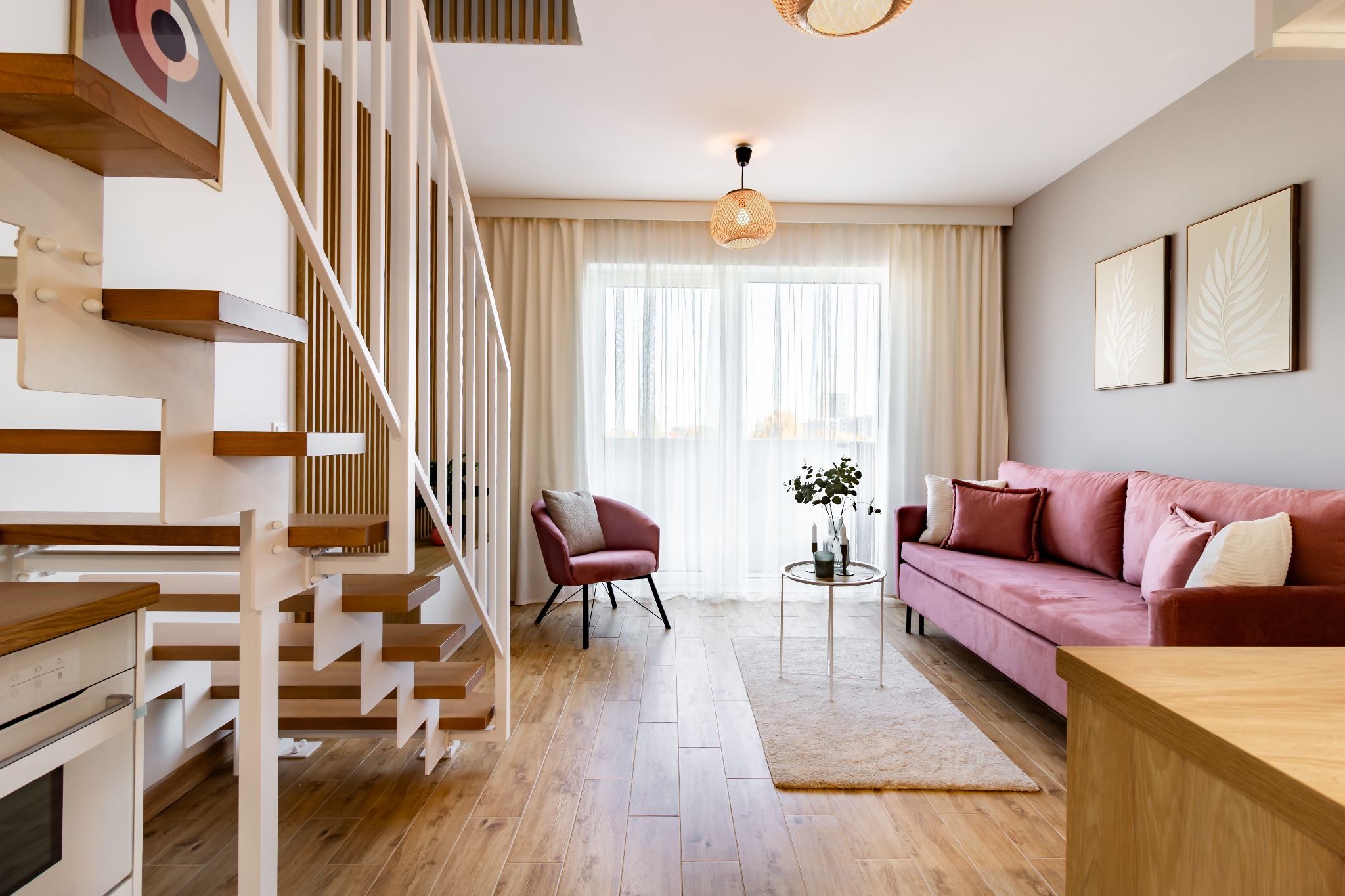 Jasne wnętrze z drewnianymi schodami, różową sofą i fotelami. Naturalne światło wpadające przez okno podkreśla przytulny charakter aranżacji.