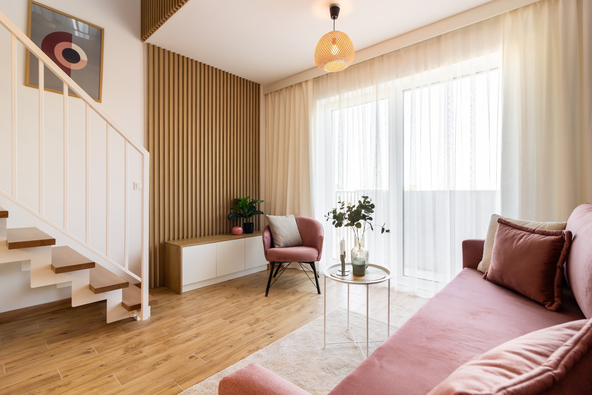 Jasne wnętrze z drewnianymi akcentami: schody, lamela na ścianie, podłoga. Różowa sofa i fotel, okrągły stolik. Minimalistyczny design, naturalne światło.
