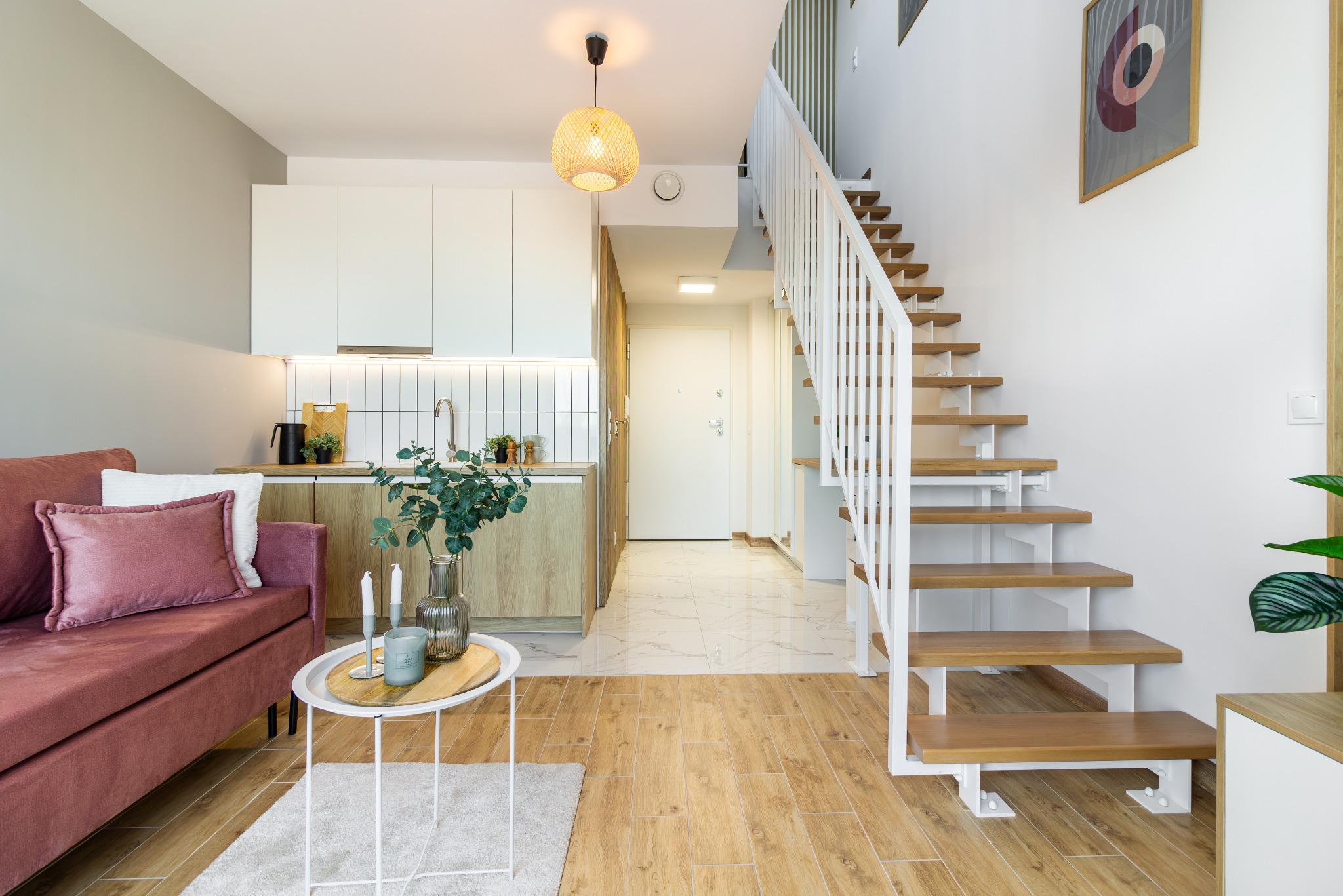 Jasne wnętrze mieszkania z drewnianymi schodami, różową sofą i okrągłym stolikiem kawowym. Minimalistyczna kuchnia w tle, całość w neutralnej kolorystyce.