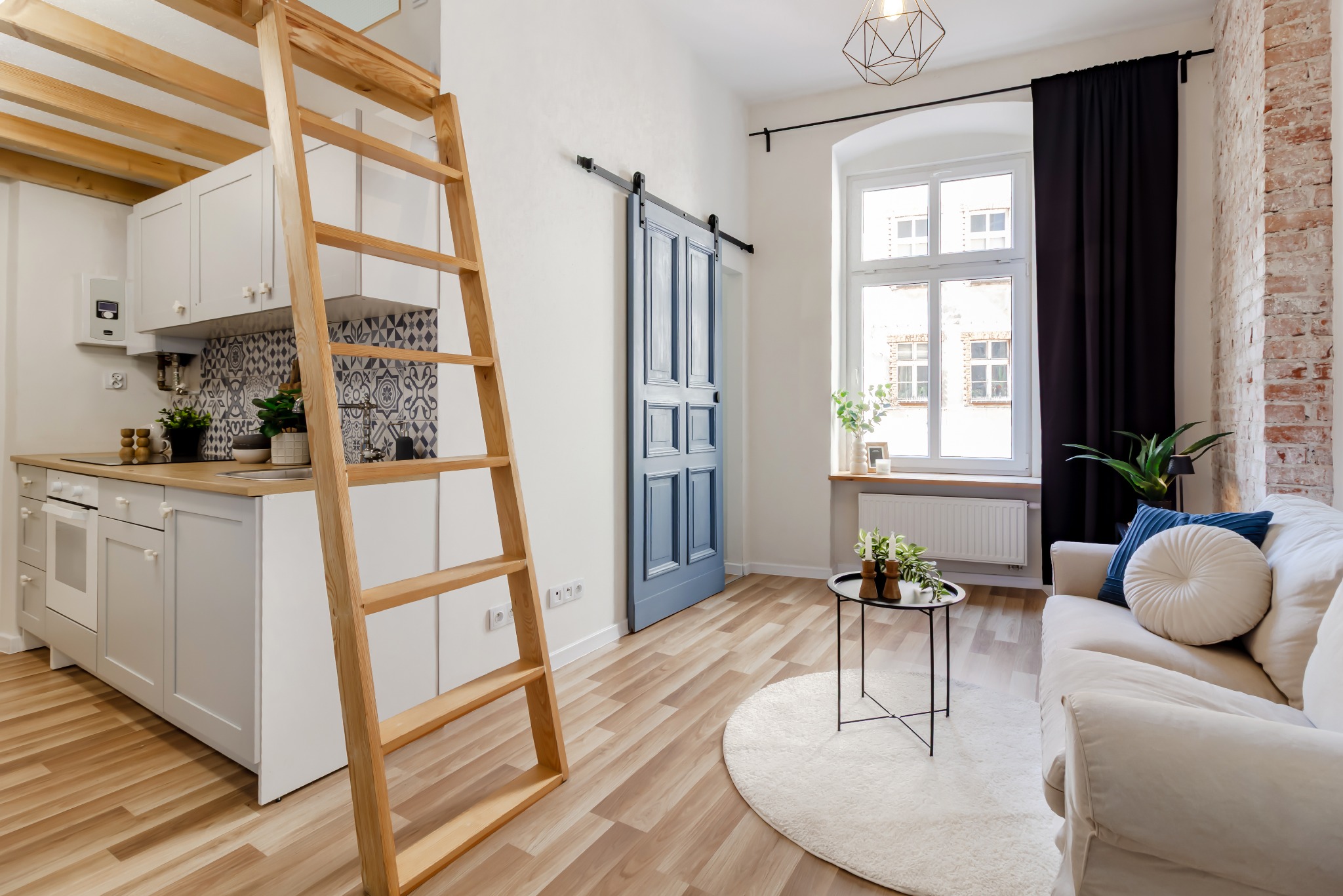 Jasne wnętrze mieszkania z antresolą: drewniana drabina, biała kuchnia z wzorzystymi płytkami, błękitne drzwi przesuwne i sofa z poduszkami. Parkiet w ciepłym odcieniu.