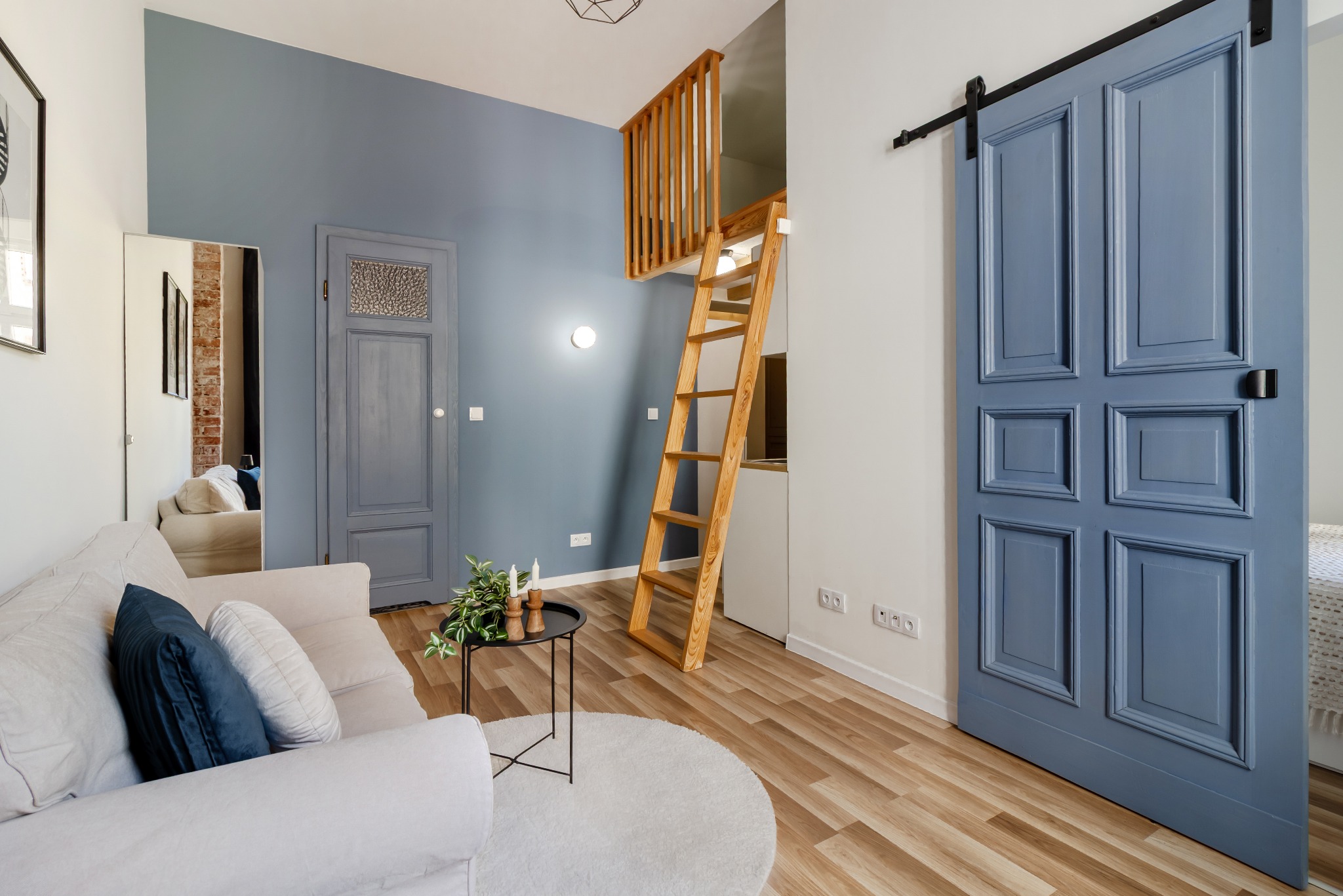 Jasne wnętrze mieszkania z antresolą i drewnianą drabiną. Niebieska ściana, sofa z poduszkami, okrągły dywan i przesuwne drzwi w kolorze niebieskim.