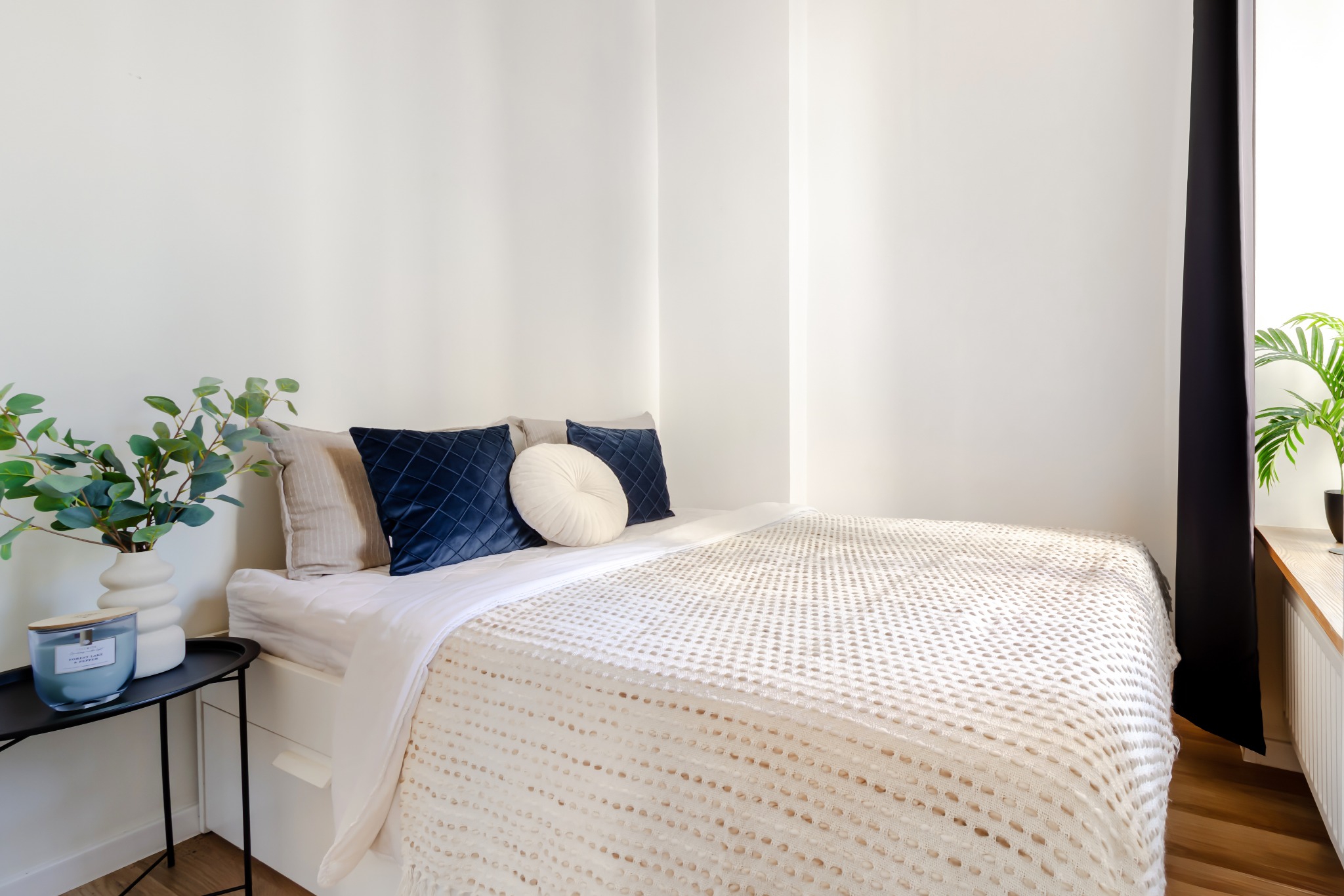 Jasna sypialnia w Kiełczowie: łóżko z dekoracyjnymi poduszkami, stolik z rośliną i świecą. Minimalistyczny design, naturalne światło, przytulna atmosfera.