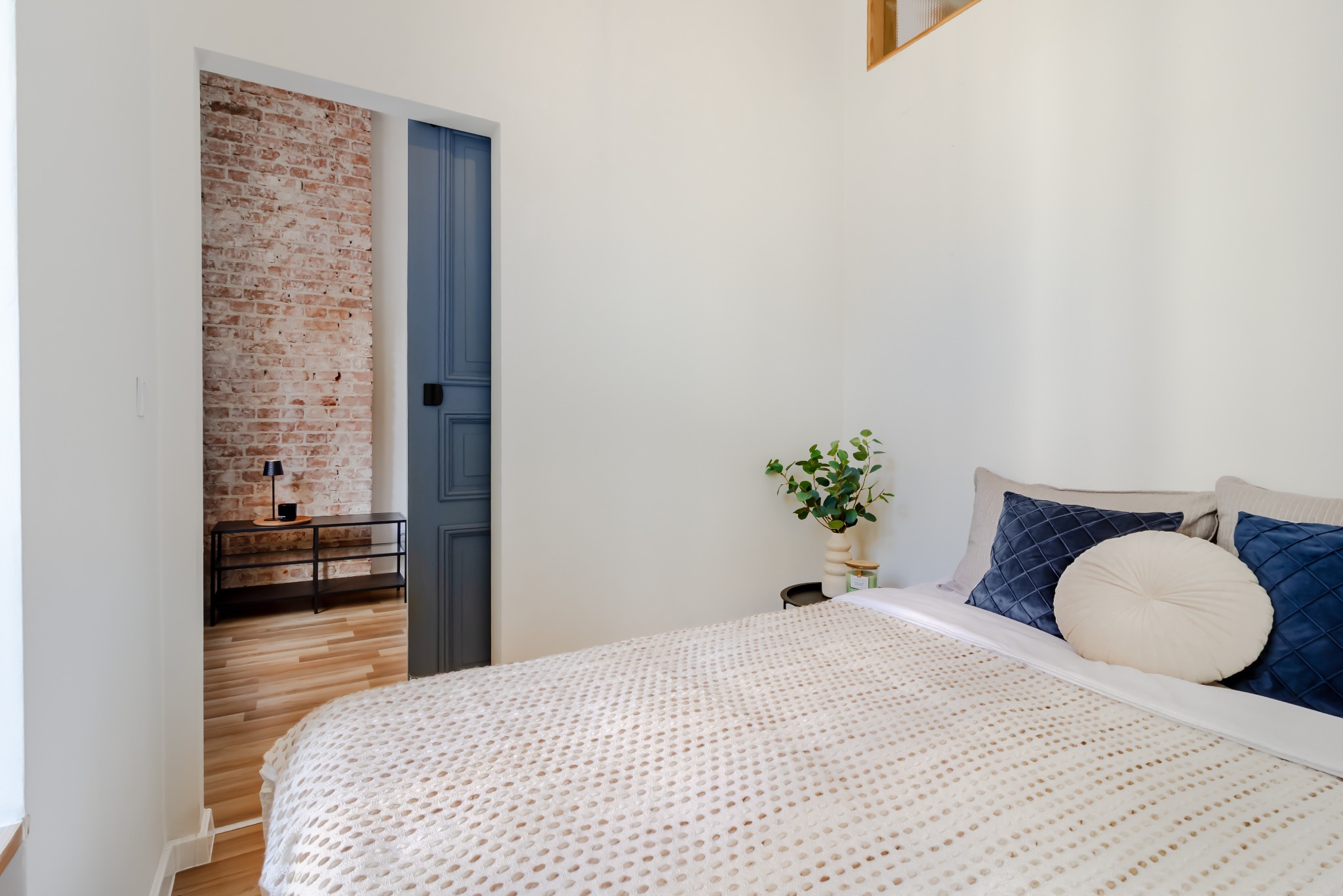 Jasna sypialnia z ceglaną ścianą w tle, niebieskie drzwi i beżowa narzuta z poduszkami. Minimalistyczny wystrój, roślina w wazonie na stoliku nocnym.