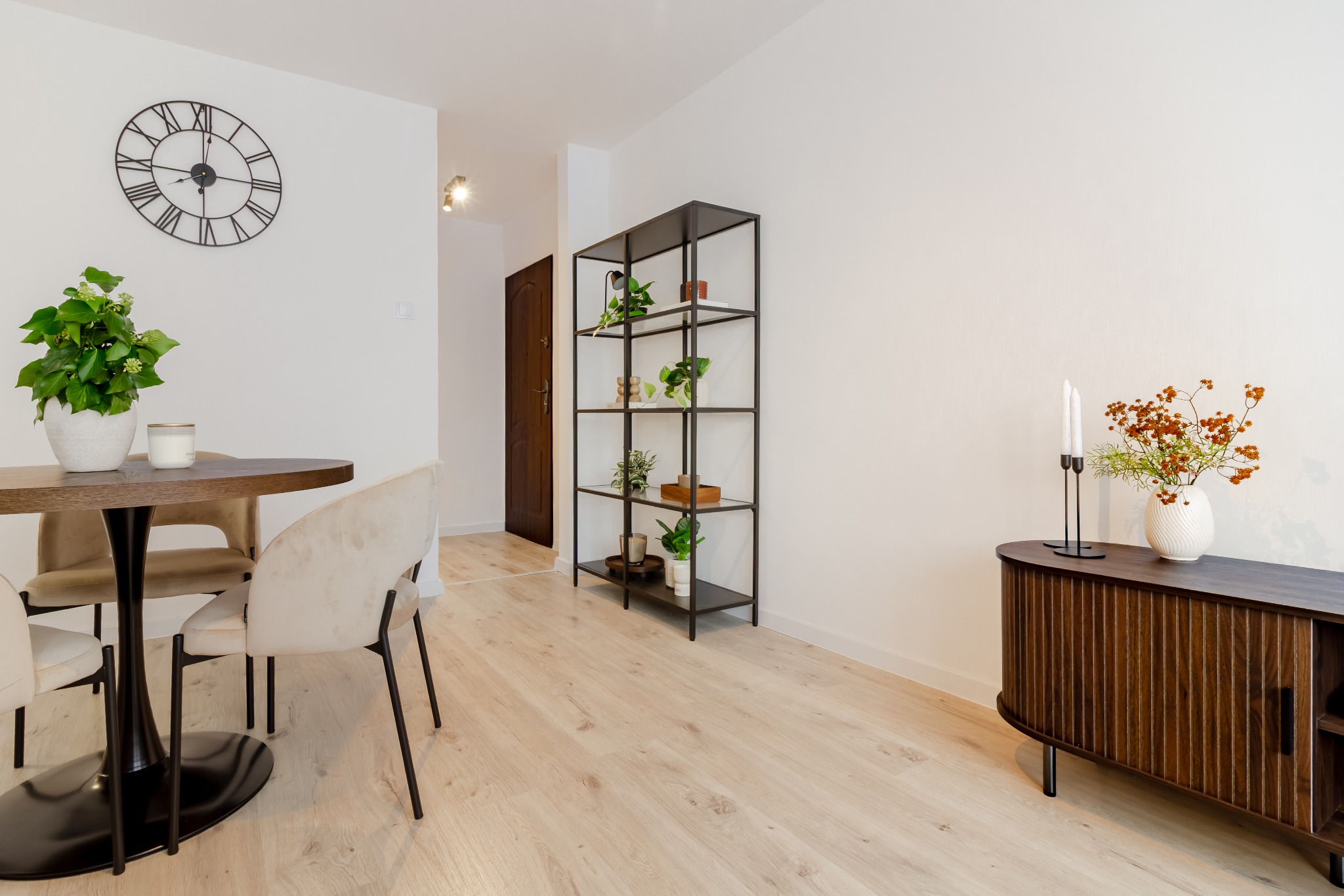Jasne wnętrze z okrągłym stołem, krzesłami w odcieniach beżu, metalowym regałem z roślinami i komodą z dekoracjami. Na ścianie widoczny zegar.