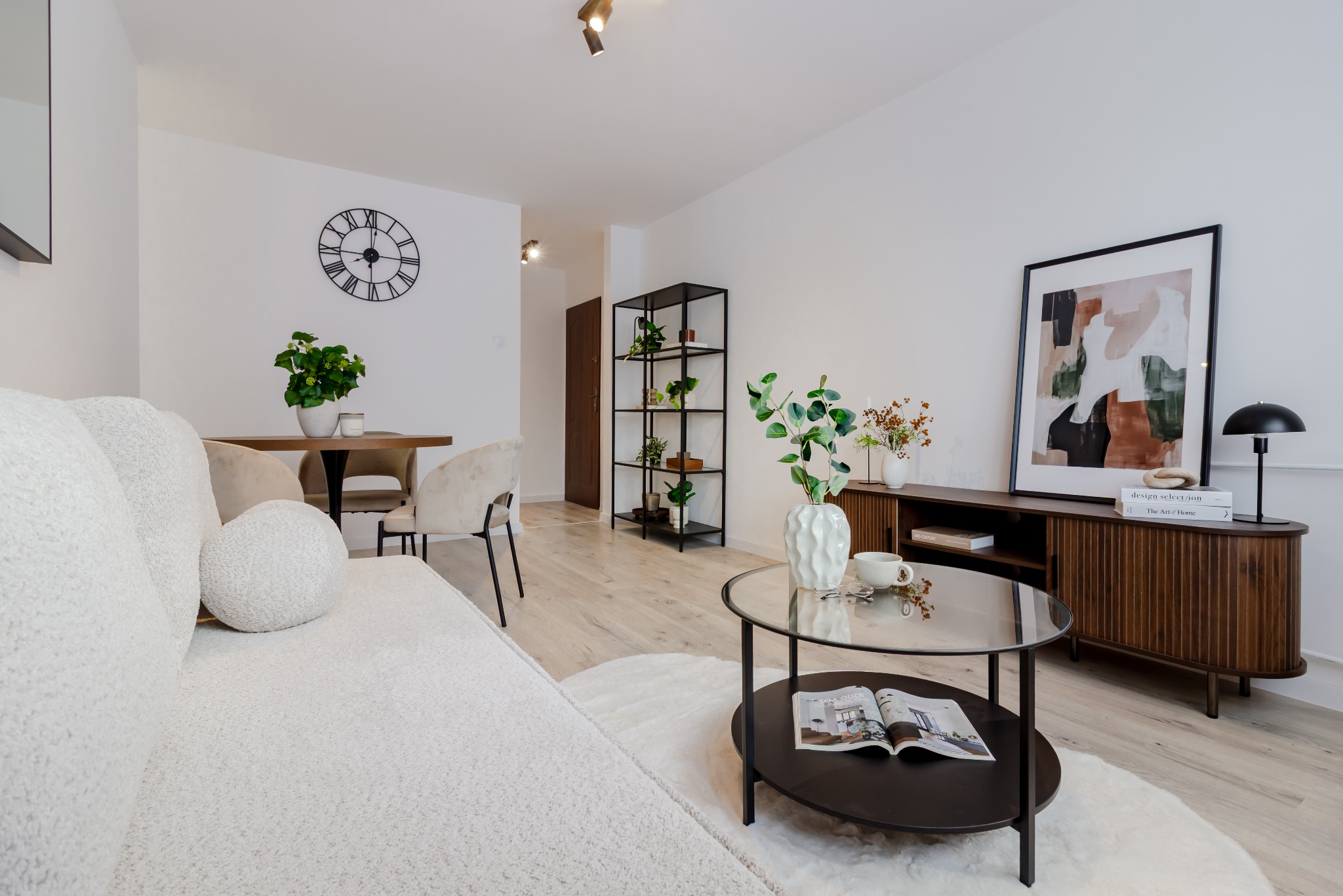 Jasne wnętrze salonu z elementami dekoracyjnymi: sofa, stolik kawowy, regał z roślinami, zegar na ścianie. Neutralna kolorystyka, minimalistyczny styl.