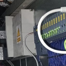 MARCIN WYRWA - Panel sterowania z modułami PLC, okablowaniem i złączami Ethernet. Widoczne oznaczenia ostrzegawcze na obudowach. Instalacja w szarej szafie sterowniczej.