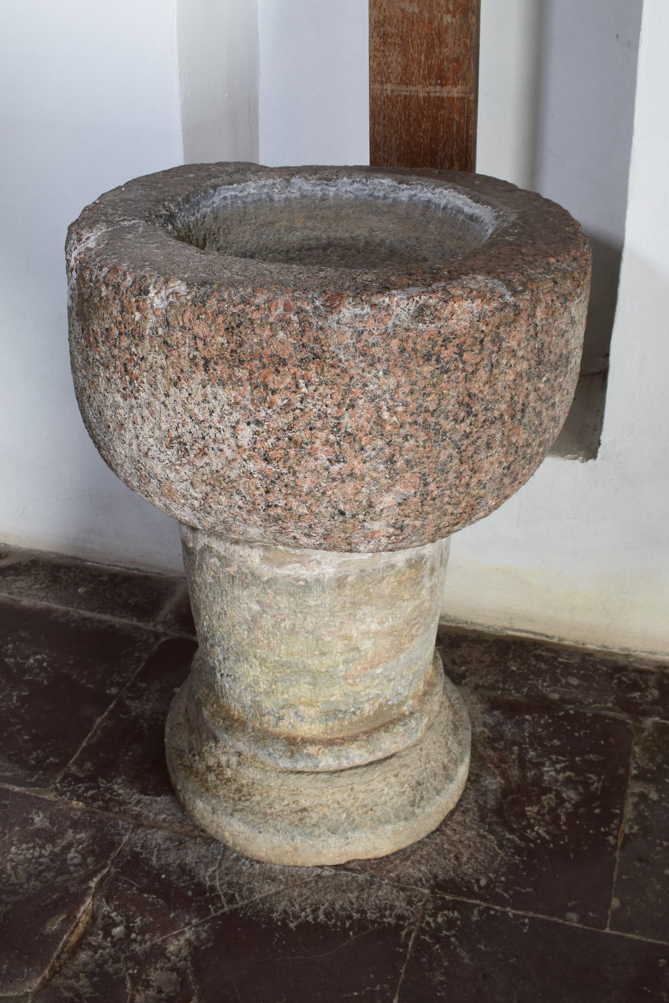 Kamienna chrzcielnica z misą z różowego granitu, osadzona na cylindrycznej podstawie, na tle białej ściany i ciemnej, marmurkowej posadzki w kościele.