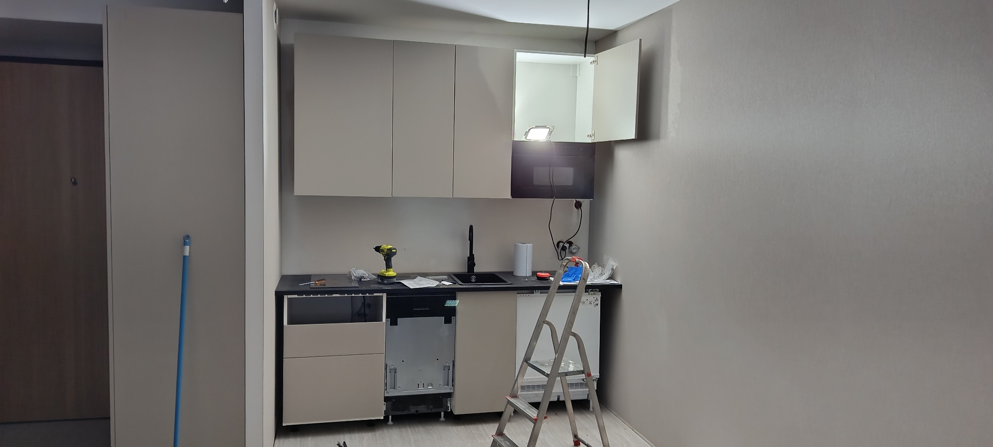 Kuchnia w trakcie montażu: widoczne szafki wiszące, częściowo zamontowane szafki dolne, drabina i narzędzia. Oświetlenie górne podkreśla otwartą szafkę.