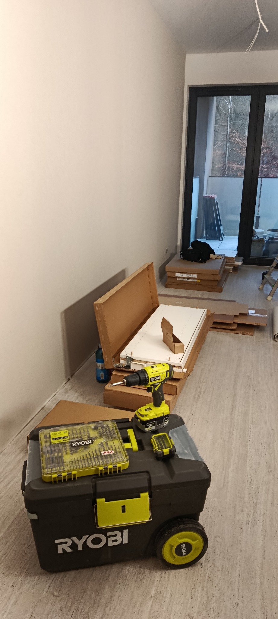 Rozpakowane elementy mebli Ryobi, skrzynka narzędziowa i wkrętarka akumulatorowa Ryobi na podłodze w jasnym pokoju z wyjściem na balkon.