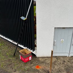 Geox Usługi Geodezyjne Rafał Gładysiak - Pomiar geodezyjny w terenie: tyczka z tabletem, skrzynka narzędziowa i młot obok ogrodzenia i ściany budynku. Oznaczenia pomiarowe na ziemi.