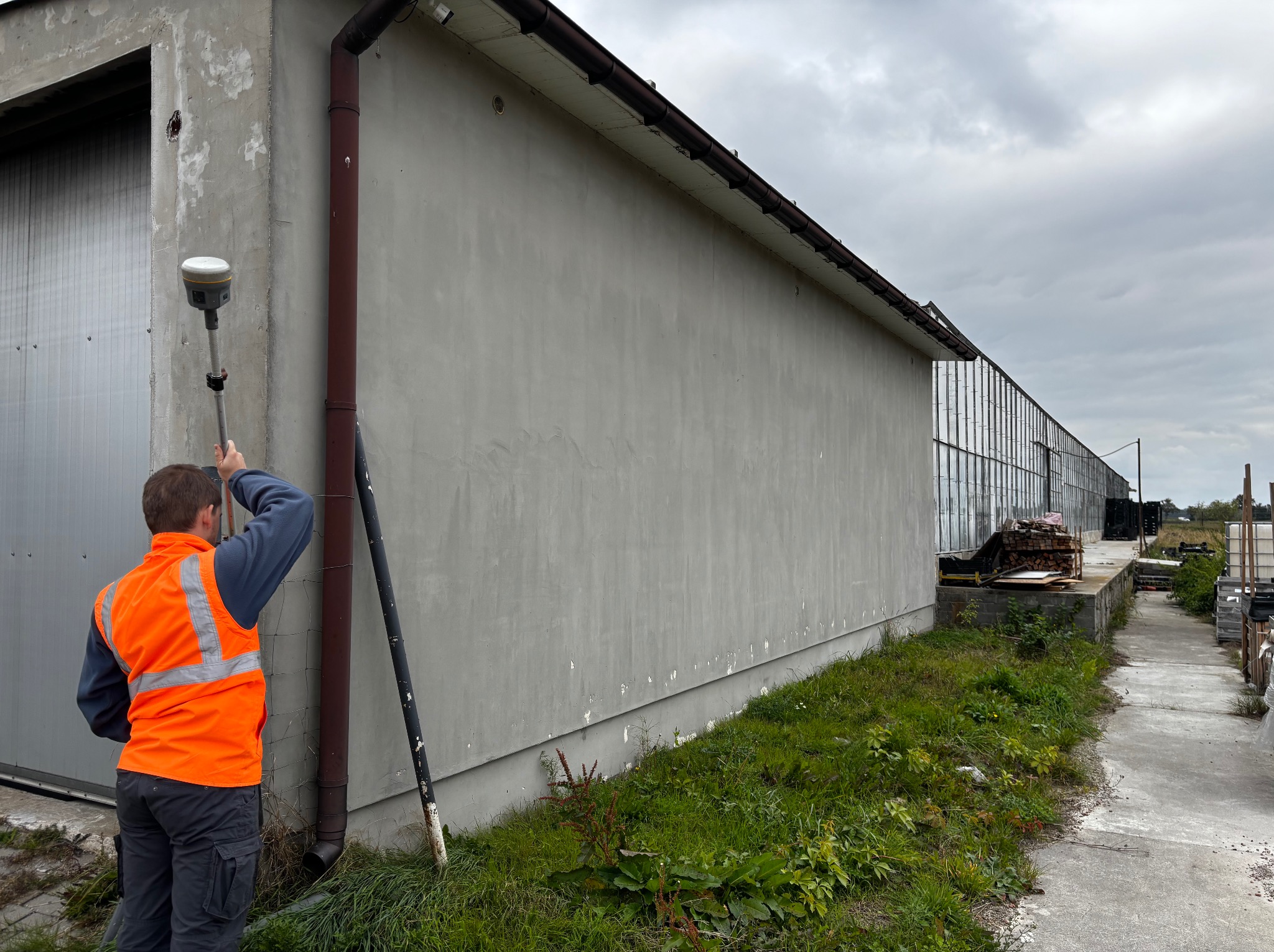 Pomiar budynku: osoba w kamizelce mierzy ścianę budynku za pomocą specjalistycznego sprzętu pomiarowego. Widoczne elementy konstrukcyjne i otoczenie.