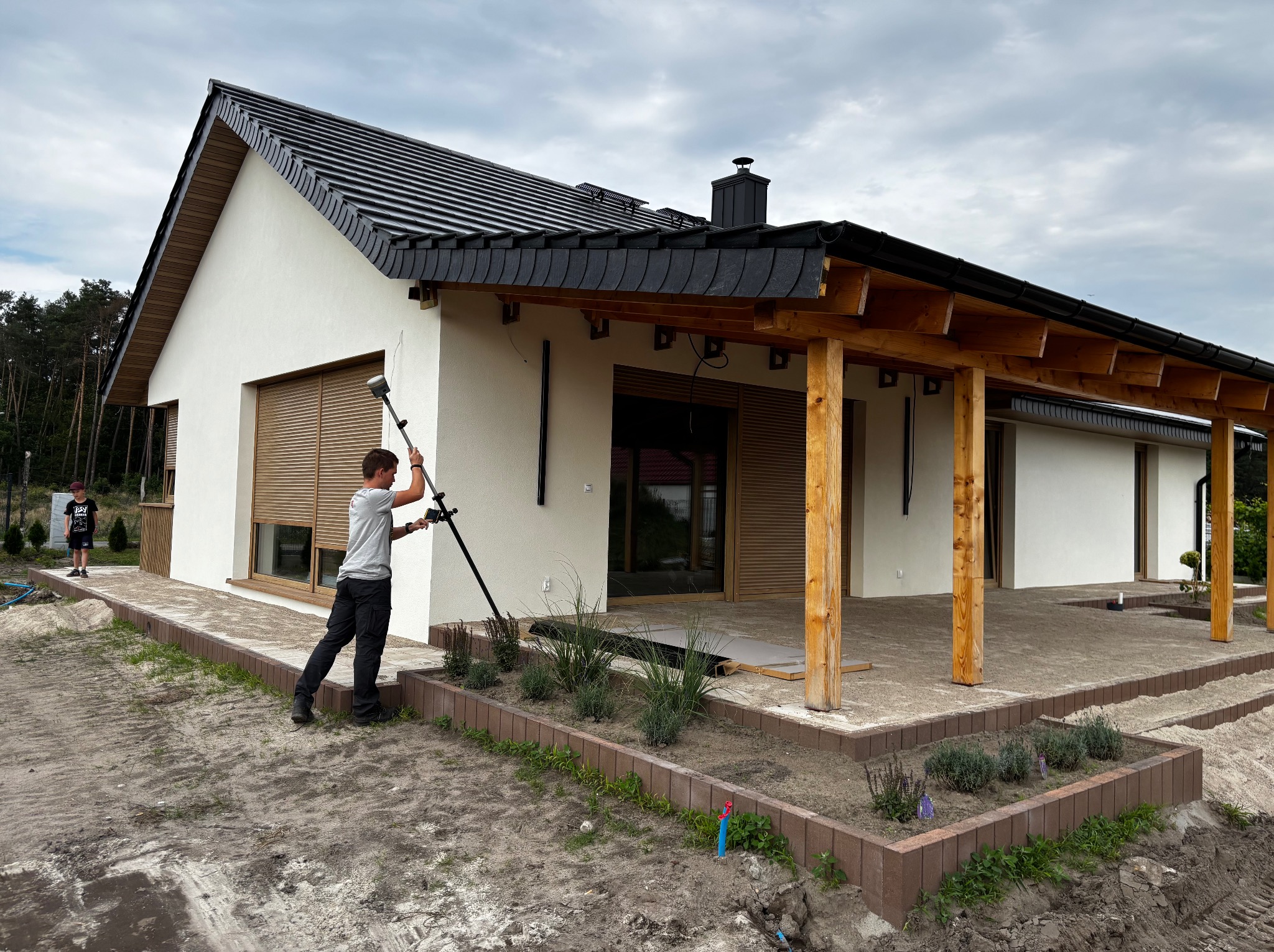 Geodeta mierzy elewację nowoczesnego domu z czarnym dachem i drewnianymi elementami. Widoczne rabaty kwiatowe i częściowo ułożona kostka brukowa.