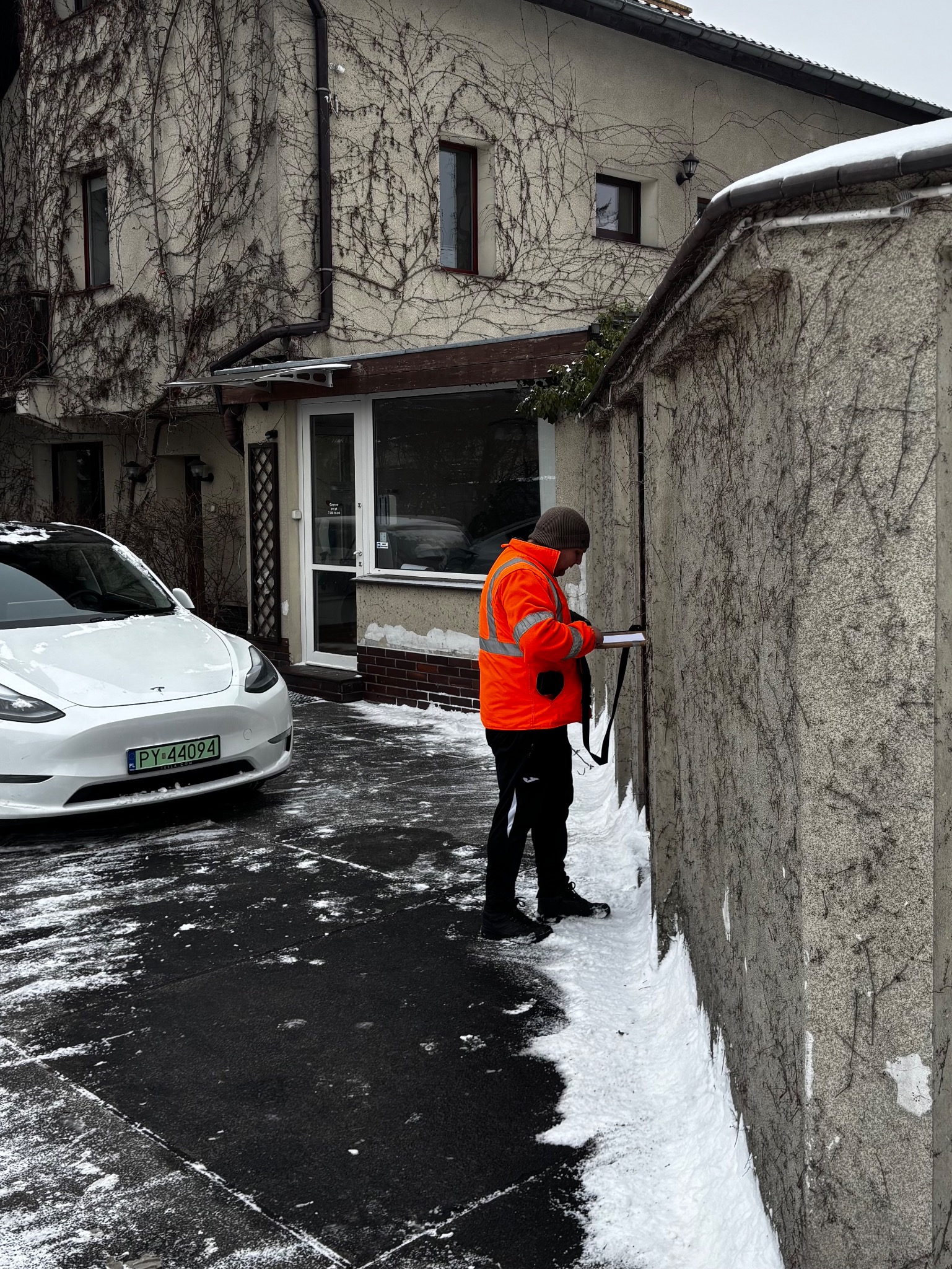 Pracownik w pomarańczowej kurtce, z dokumentami przy murze porośniętym bluszczem. W tle biały samochód i budynek. Zimowa sceneria.