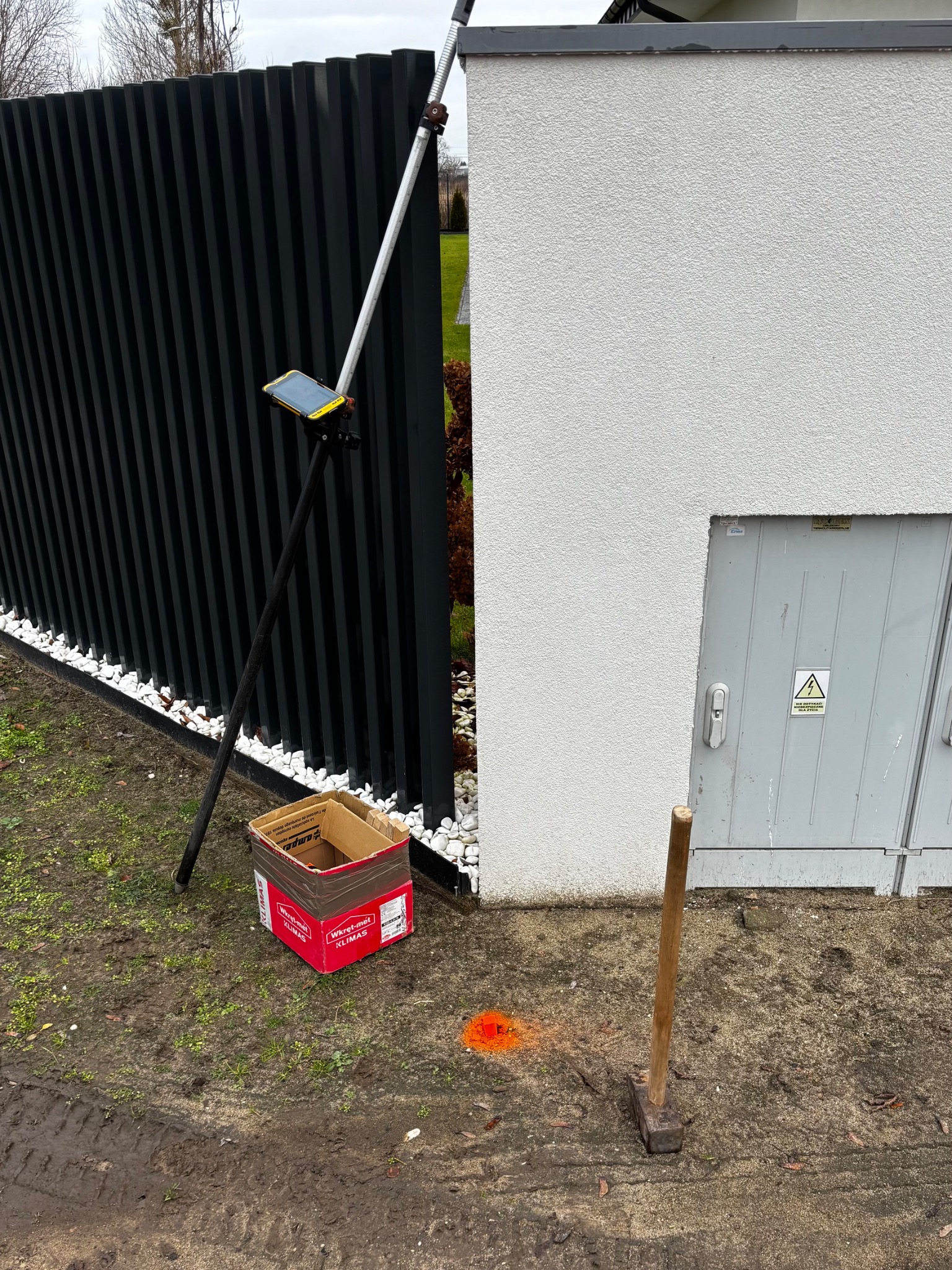 Pomiar geodezyjny w terenie: tyczka z tabletem, skrzynka narzędziowa i młot obok ogrodzenia i ściany budynku. Oznaczenia pomiarowe na ziemi.