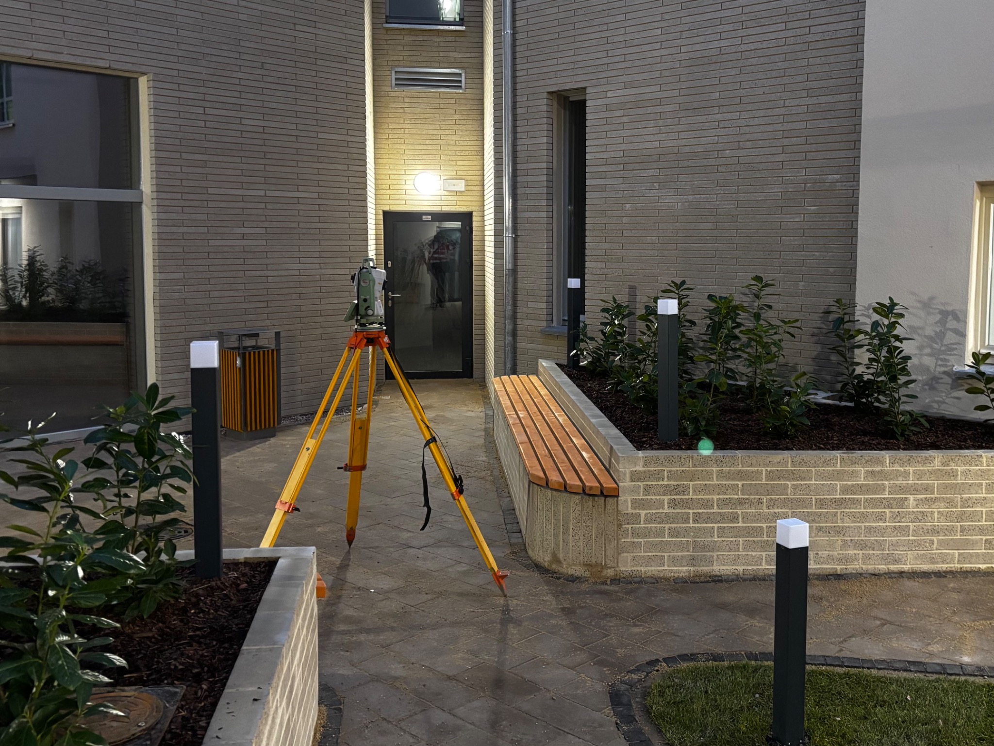 Tachimetr na statywie ustawiony na brukowanym dziedzińcu między budynkami z cegły, oświetlony lampami. W tle widoczne drzwi wejściowe.