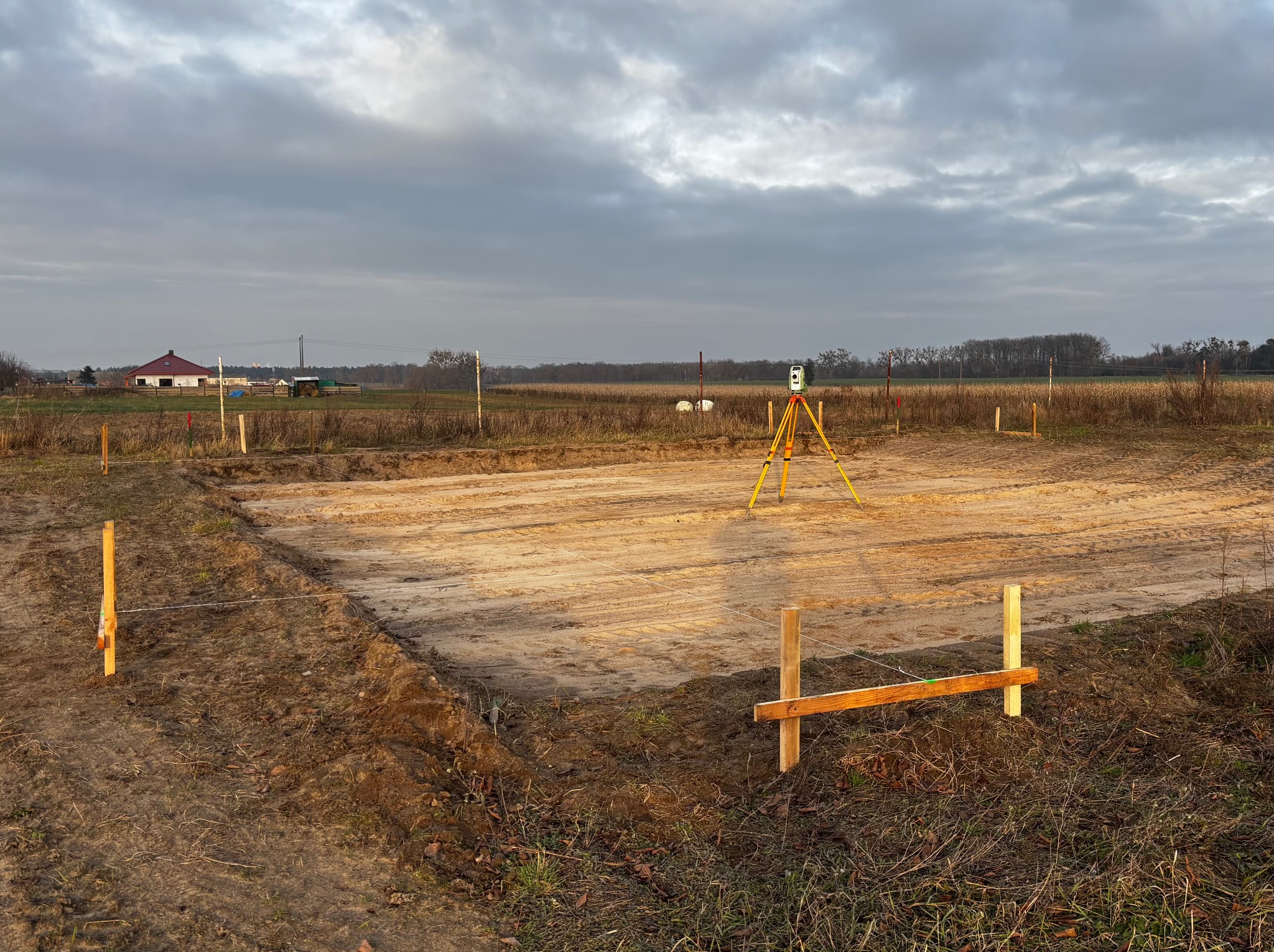 Wytyczanie fundamentów pod budowę w Kaźmierzu. Żółty niwelator na trójnogu, drewniane paliki z linkami, przygotowany teren pod budowę, pochmurne niebo.