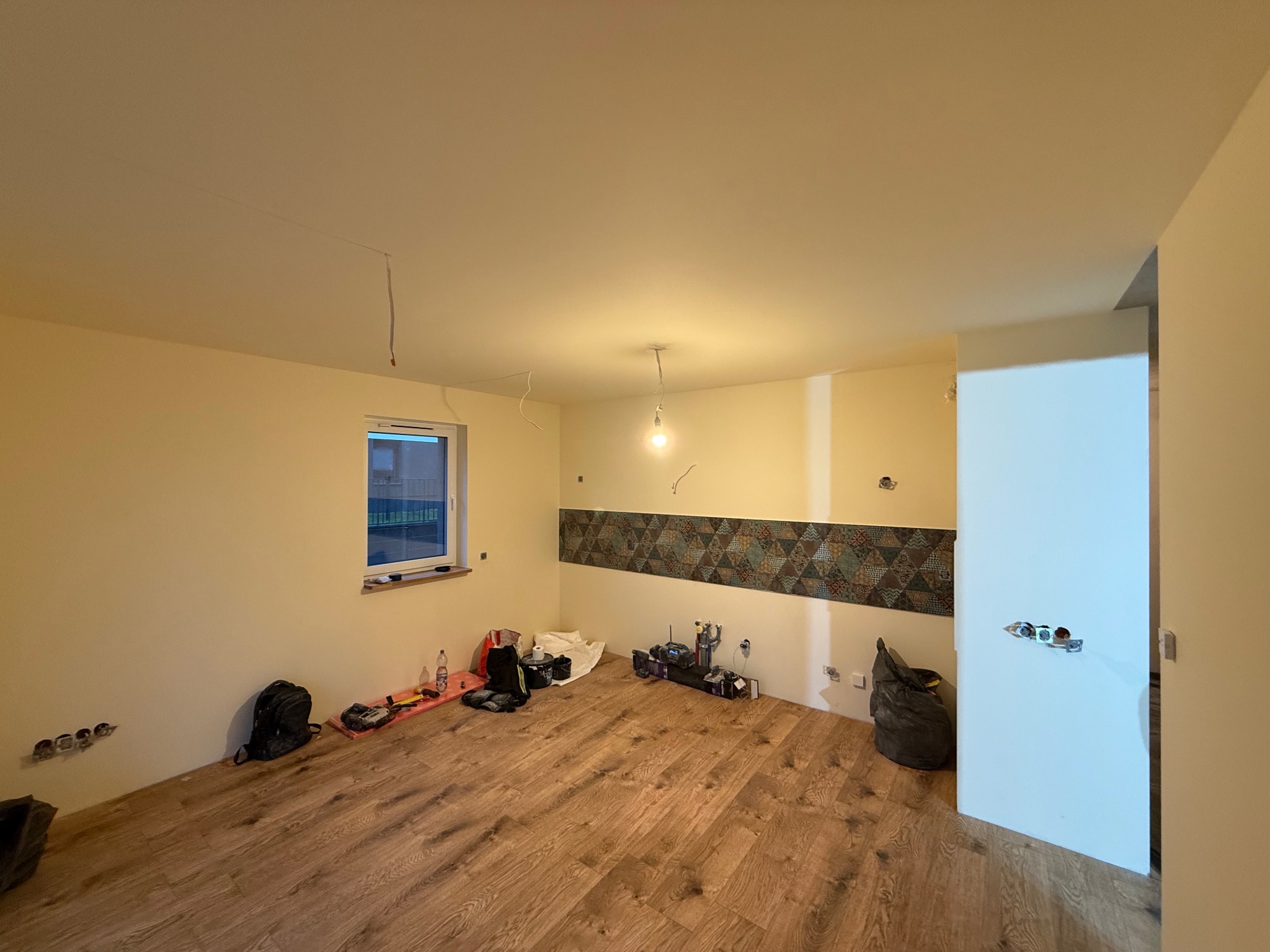 Pokój w trakcie remontu: drewniana podłoga, ściany w jasnym odcieniu, widoczne narzędzia, niedokończone gniazdka elektryczne i fragment dekoracyjnej listwy na ścianie.