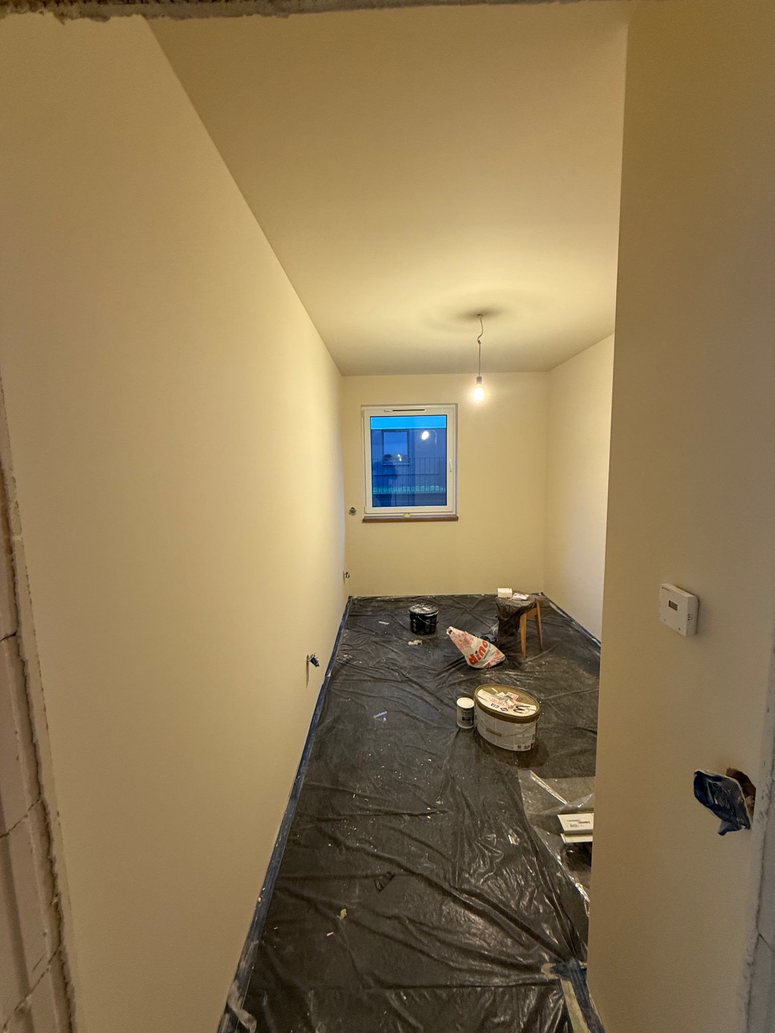 Pomieszczenie po malowaniu ścian na jasny odcień, zabezpieczone folią, widoczne puszki z farbą i krzesło. Ściany gładkie, świeżo pomalowane, widok z progu.