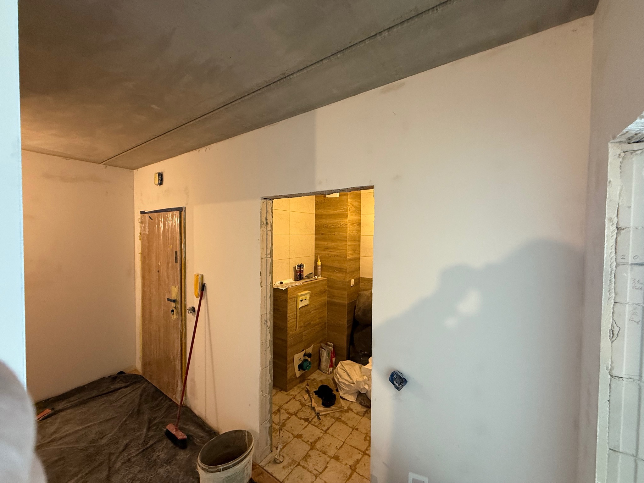 Wnętrze w trakcie remontu: surowy betonowy sufit, świeżo otynkowane ściany, widoczne przygotowania do dalszych prac wykończeniowych, otwarte przejście do wykafelkowanej łazienki.