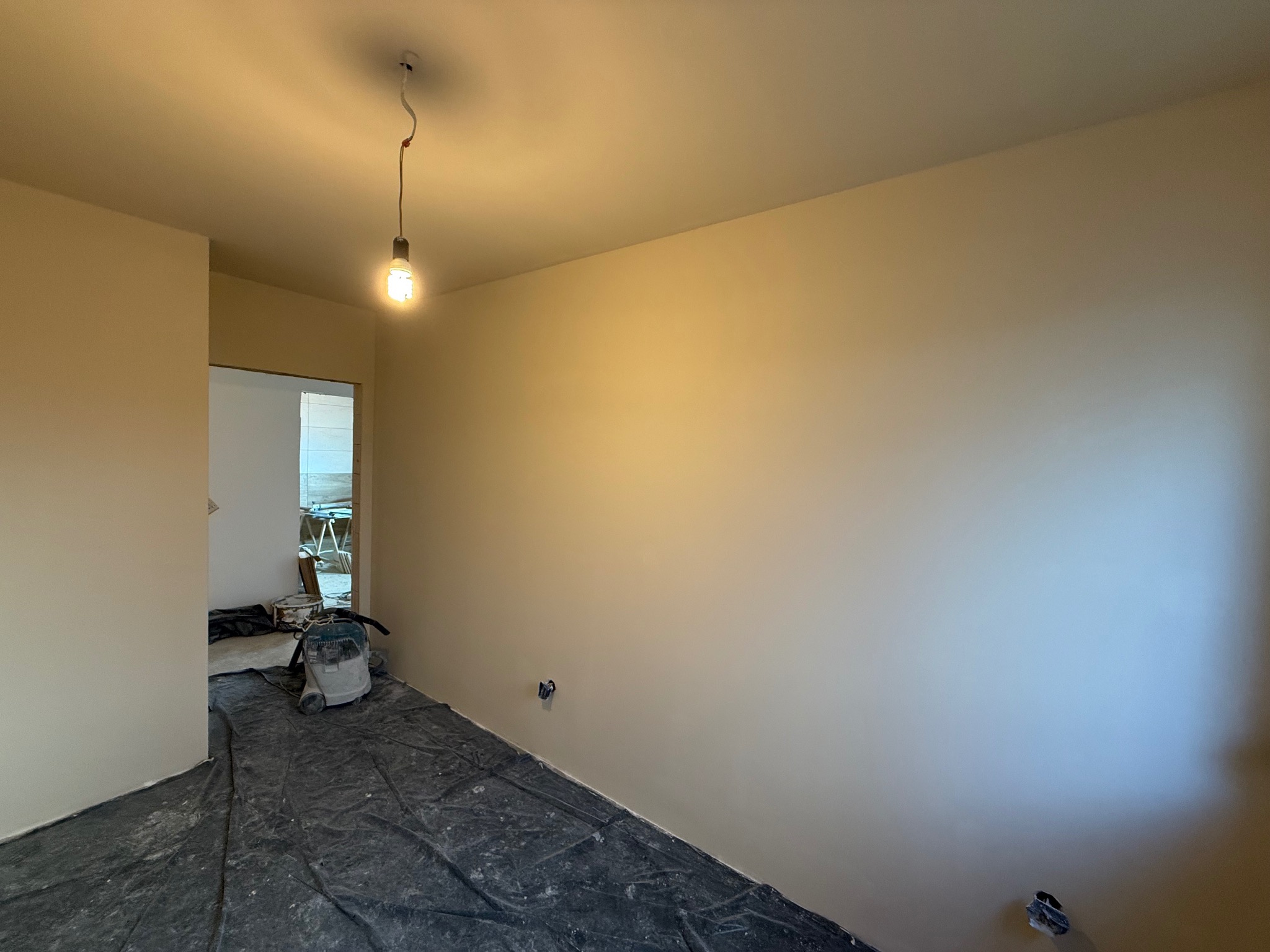 Świeżo pomalowane ściany w pokoju z podłogą zabezpieczoną folią malarską. Widoczny odkurzacz przemysłowy i wisząca żarówka. Gładkie, beżowe ściany po malowaniu.