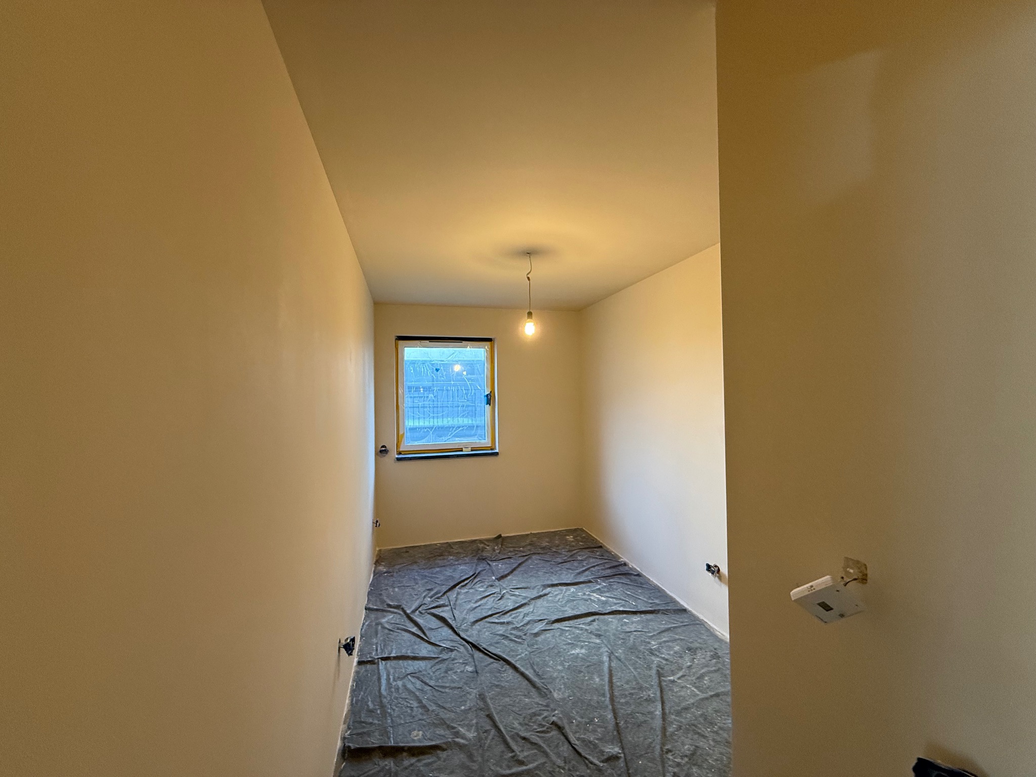 Pokój po szpachlowaniu, przygotowany do malowania. Podłoga zabezpieczona folią, ściany gładkie, w oknie widoczne zabezpieczenia. Wnętrze oświetlone wiszącą żarówką.