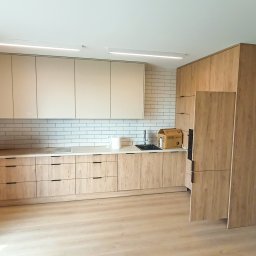 Warmińskie Domy PATRYK GICZEWSKI - Jasna kuchnia z drewnianymi szafkami i białymi płytkami na ścianie. Widoczny zlew, blat i oświetlenie sufitowe. Minimalistyczny design, świeżo po remoncie.