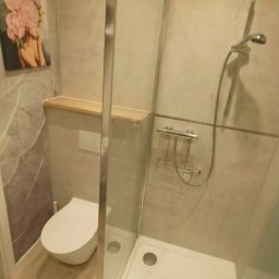 Warmińskie Domy - Nowoczesna łazienka z kabiną prysznicową typu walk-in, toaletą podwieszaną i półką z jasnego drewna. Na ścianie grafika z motywem kwiatowym.
